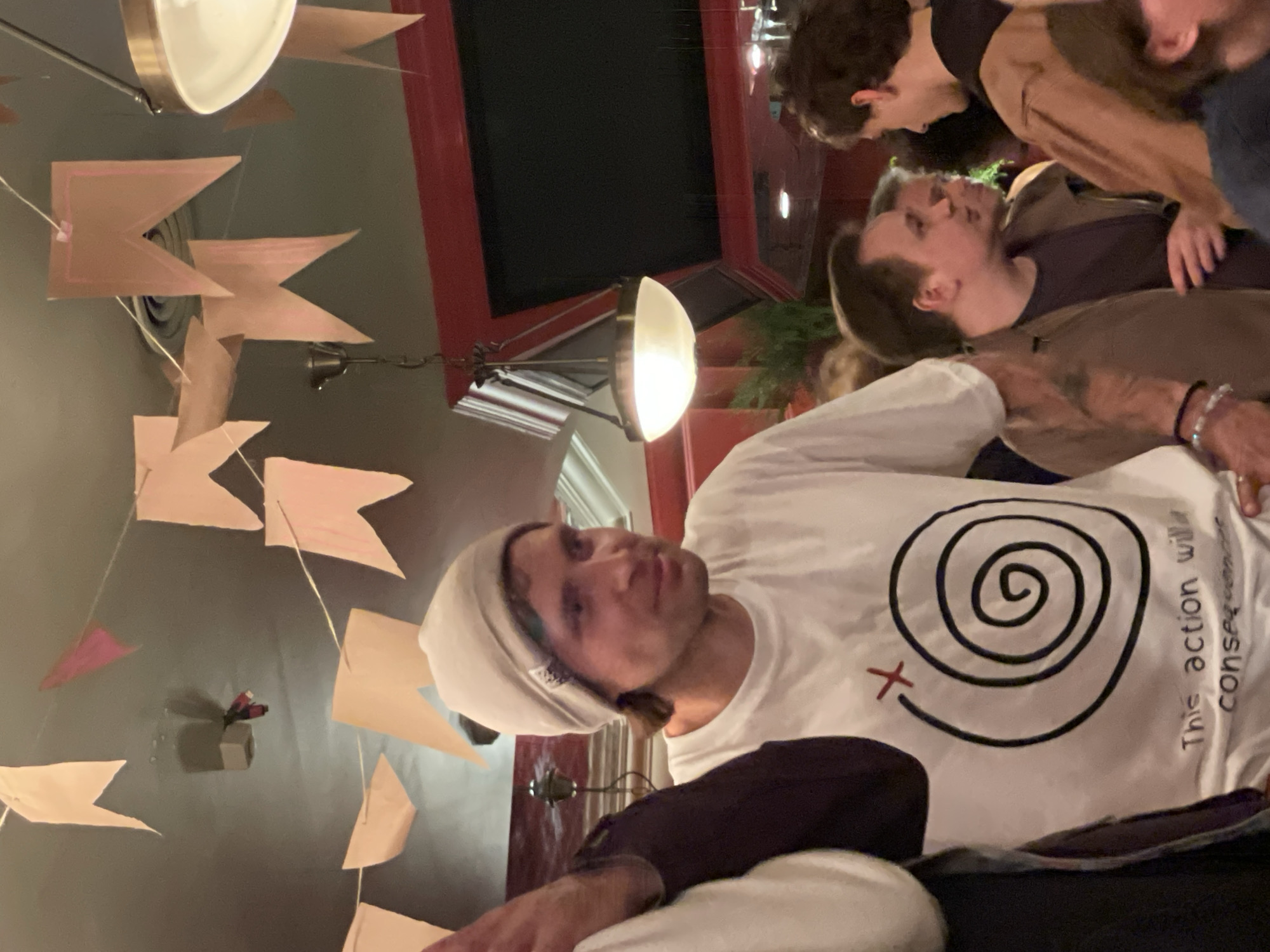 A low-angle shot shows a man wearing a white beanie and a white t-shirt with a black spiral and red "X" design, as well as text reading "This action will consequences." He is positioned in front of several people standing close together. The background includes what appears to be party decorations consisting of numerous tan pennant flags. There is a lamp hanging in the background with a curved white shade. Red architectural molding surrounds a dark, rectangular space, possibly a window or screen. The photo was likely taken indoors. It is not possible to discern any details regarding the location as Moscow, Russia.