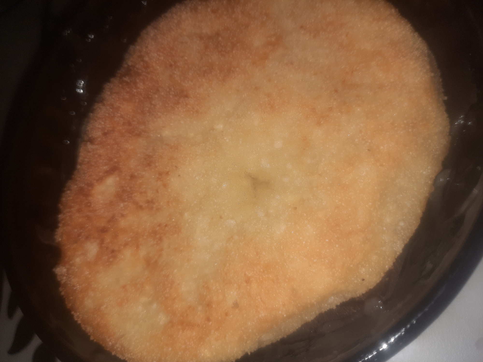 An overhead shot of a single, golden-brown, round, fried pastry sits on a dark, reflective surface, possibly a plate or pan. The pastry has a slightly irregular shape and its surface is textured with a fine, granular appearance. There are some darker brown, slightly charred areas around the edges and in some spots on the surface, indicating it has been fried. The background is dark and out of focus, with some bright highlights suggesting it is a reflective material. The lighting appears to be artificial and directly above, casting a soft glow on the pastry. There are no people or other objects visible in the image. The location in the city of Shklow, Belarus is not discernible from the image.