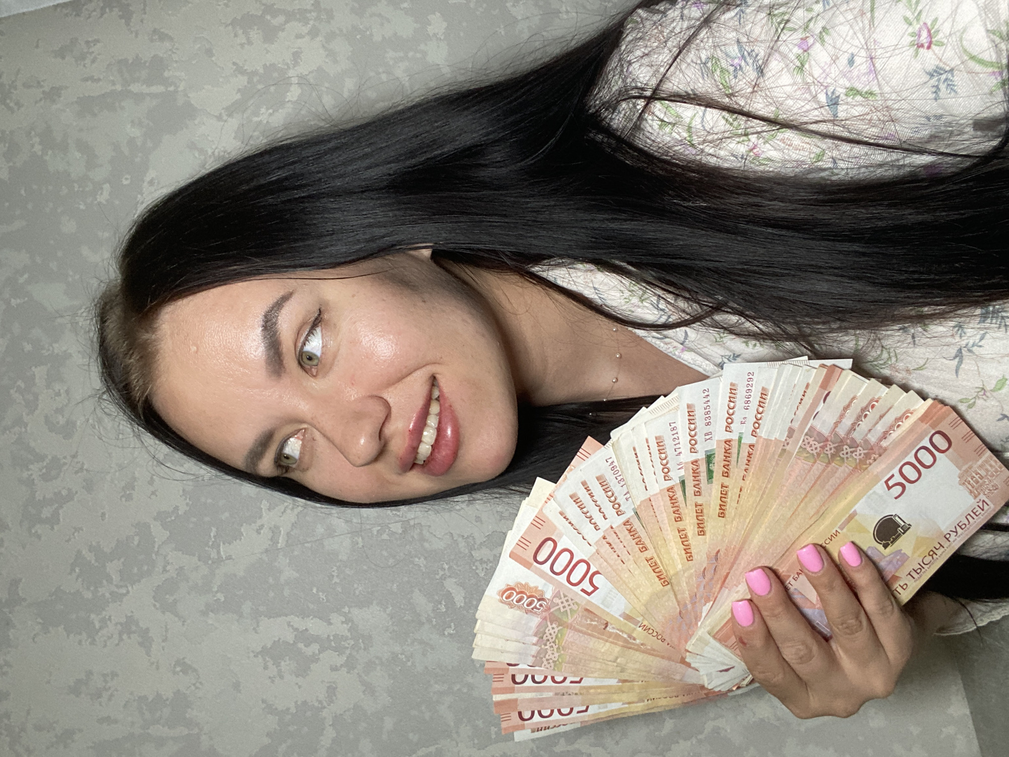 The image shows a young woman with long, dark hair and light-colored eyes. She is holding a fan of Russian 5000 ruble banknotes. Her nails are painted pink. She is wearing a light-colored shirt with a floral pattern. The background is a textured gray wall. There is no indication of the specific location in the city of Chaikovsky, Russia.