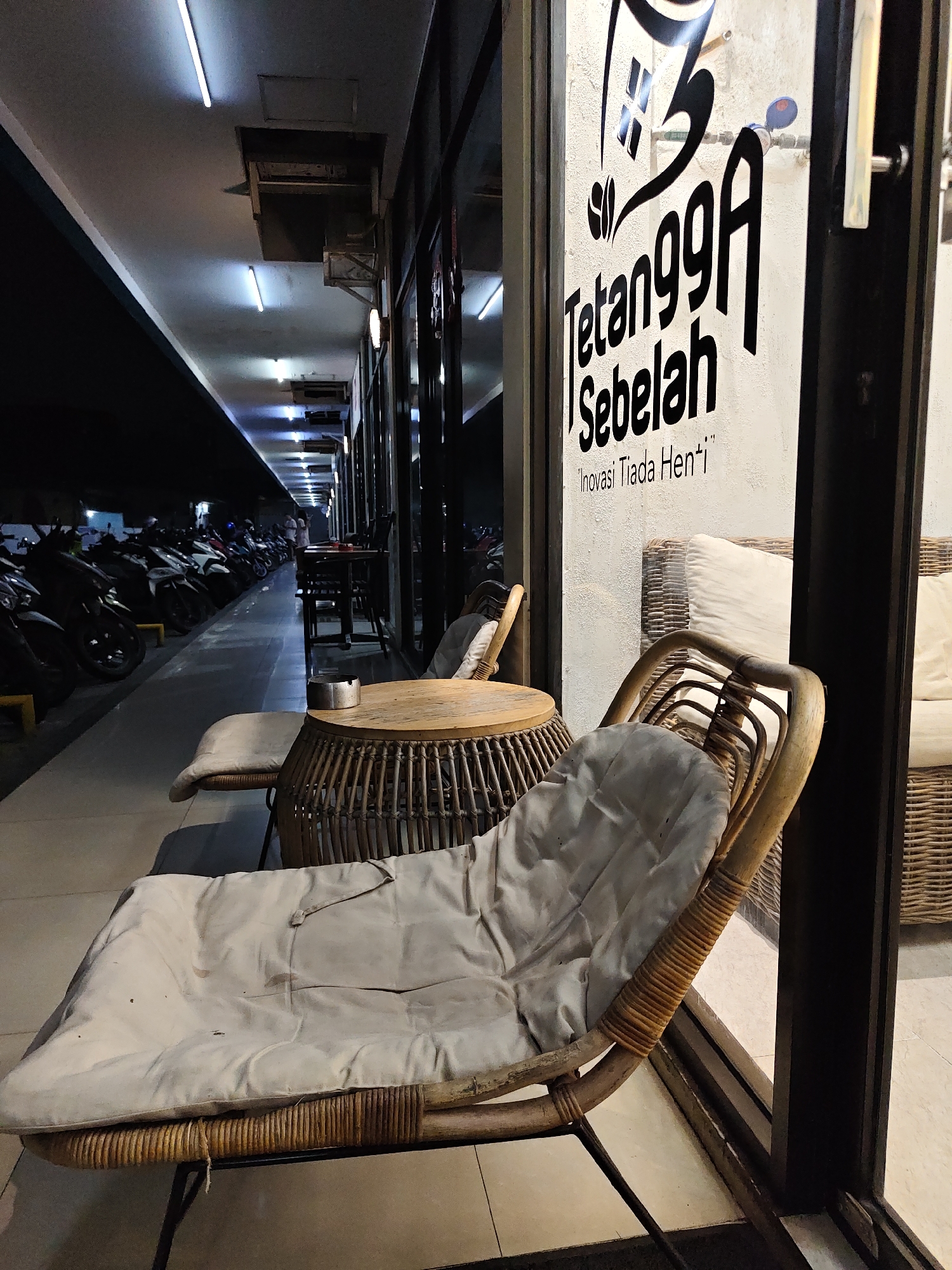 The image depicts an outdoor seating area of a cafe at night, located in North Jakarta, Indonesia. The cafe's facade is visible, featuring a large window with a logo that reads "Tetangga Sebelah" and the tagline "Inovasi Tiada Henti". Inside the cafe, wicker furniture is arranged, including a sofa with white cushions. Outside, a round wicker coffee table with an ashtray sits in front of a wicker lounge chair with a light-colored cushion. Further back, a row of motorcycles is parked along a walkway, illuminated by overhead fluorescent lights. The environment suggests a commercial district or parking area adjacent to the cafe.