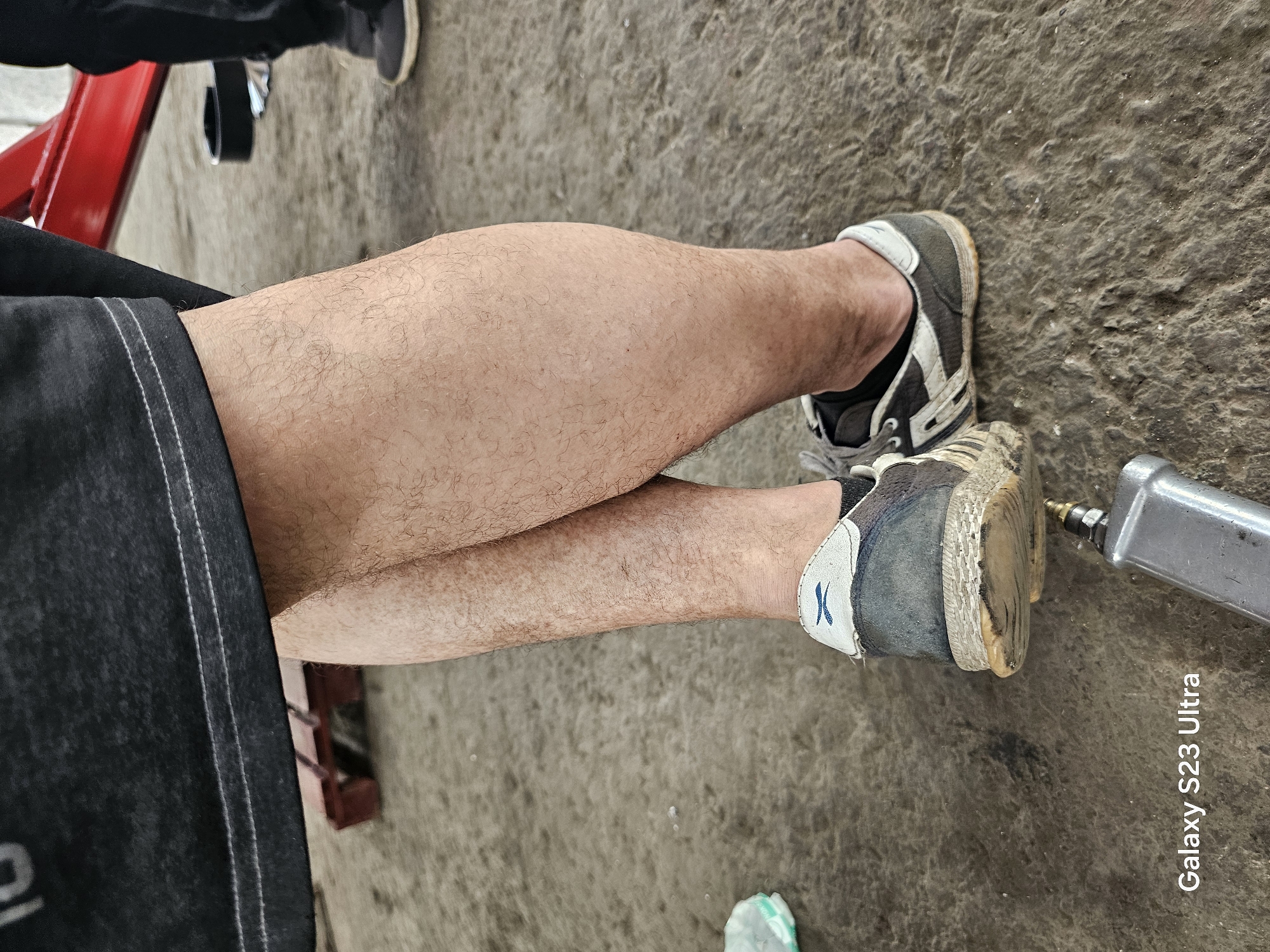 The image presents a ground-level perspective of crossed legs and feet, likely in an outdoor, industrial or garage setting. The legs are fair-skinned and covered with fine hair. The left leg is slightly bent and positioned above the right leg. The person is wearing dark shorts with a light-colored seam. The footwear consists of worn, gray-and-white athletic shoes with visible dirt and wear on the soles and sides. To the right of the feet, a silver-colored pneumatic tool is partially visible. The background consists of a rough, textured gray surface, possibly concrete or asphalt. The bottom right corner contains the text "Galaxy S23 Ultra," suggesting the image was taken with a Samsung Galaxy S23 Ultra smartphone. The red metal structure is a mechanic's tool stand with a lift. The location is outside, but there is no location of the city of Karnobat, Bulgaria in the image.