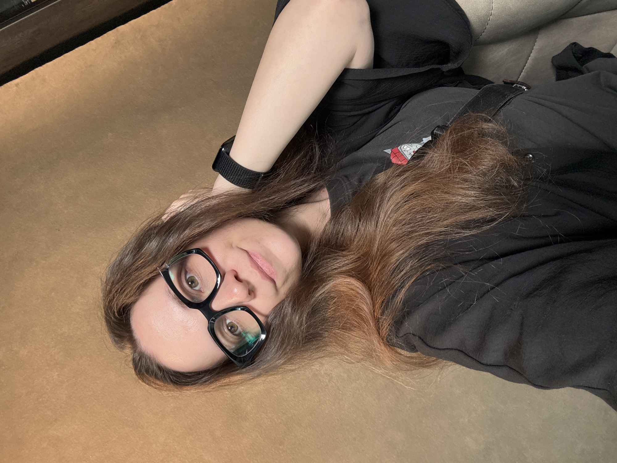 A woman with long auburn hair is lying on a brown surface. She is wearing black-rimmed glasses and a black T-shirt. A black smartwatch is visible on her left wrist. The location is unknown.