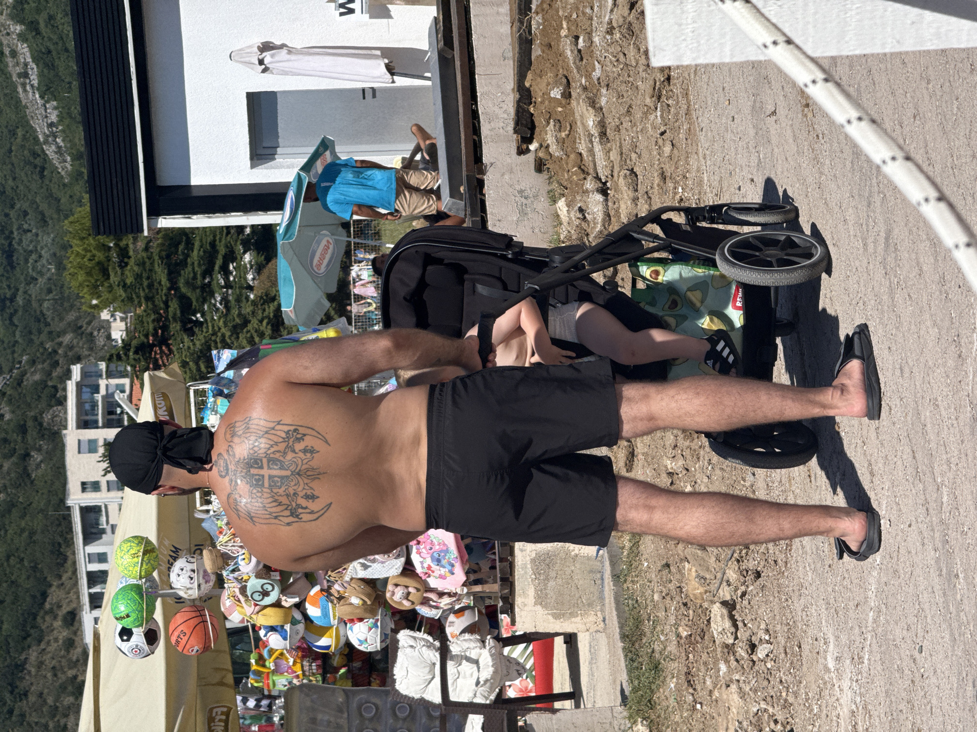 In Boreti, Montenegro, on a sunny day, a man with a large eagle tattoo on his back and wearing black shorts and a black bandana stands facing away from the camera, pushing a black stroller. Inside the stroller, a young child wearing a diaper and black sandals is visible. In the background, a stall displays numerous colorful balls and plush toys. To the right, a white building with a black roof features a closed door and a folded white umbrella. Further in the background, a man in a blue shirt and tan shorts stands near a green umbrella. The ground is a mixture of dirt and gravel, with some areas of green grass and plants. A thick white rope with black stripes runs diagonally across the lower right corner of the image.