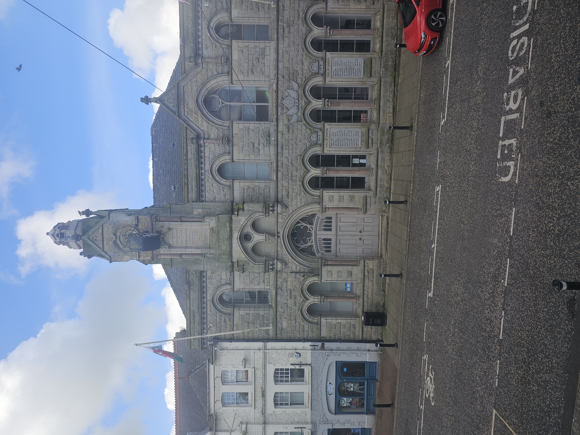 The image captures a street scene in Holywell, United Kingdom. In the foreground, there is a road marked with "DISABLED" text, indicating a parking spot for people with disabilities. A red car is parked on the side of the road.
Adjacent to the road, a building with a stone facade dominates the scene. The building's architecture is notable for its Gothic Revival style, featuring arched windows, intricate stonework, and a tall tower crowned with a spire. Plaques bearing names of historical significance are attached to the building, possibly serving as a war memorial or a "Roll of Honour".
Next to the substantial stone building, a smaller, white building with a blue storefront is visible. A Welsh flag is flying atop this smaller building. The sky above is partly cloudy with blue sky visible.