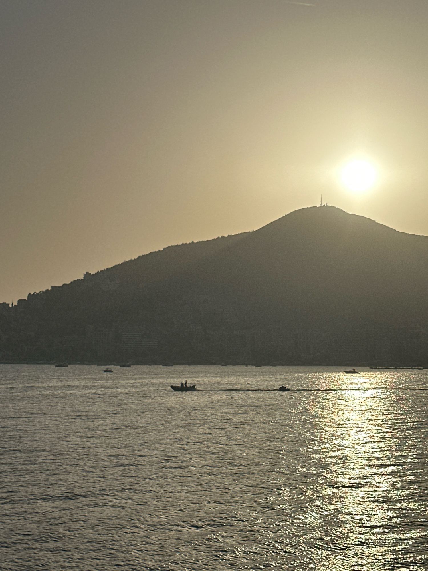 The image depicts a sunset over the Adriatic Sea in Budva, Montenegro. A large, silhouetted mountain dominates the background, with a communication tower visible at its peak. The sky is a hazy yellow, with the sun positioned high and slightly to the right, casting a bright glare. The water in the foreground is choppy, reflecting the sunlight in shimmering streaks. Several boats are scattered across the water: one larger boat with at least two people on board is in the middle ground, and a jet ski is further to the right, leaving a wake. In the distance, along the base of the mountain, there are faint indications of buildings and structures. The overall scene is one of natural beauty and recreational activity at dusk.