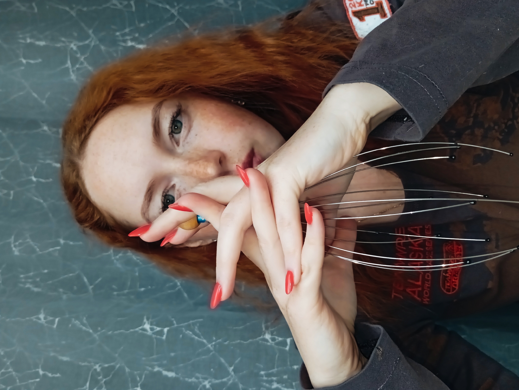 A woman with red hair and freckles is shown in a close-up shot. She is wearing a dark gray long-sleeved shirt with a patch visible on the upper right sleeve. Her fingernails are painted red. She is holding a silver head massager in her hands. The background appears to be a light blue surface with a marble-like pattern. The shirt has the words "TEAM ALASKA" on it. The location is not ascertainable, and no information indicates that the image was taken in Yurgamysh, Russia.