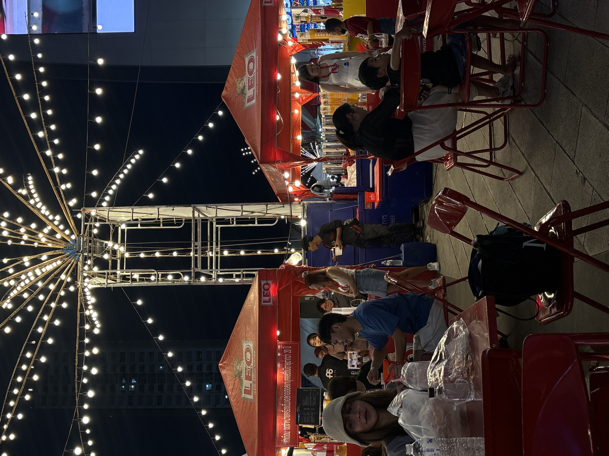 This image depicts an outdoor night market scene in Chiang Mai, Thailand, likely within the city municipality. The foreground is dominated by red tables and chairs, suggesting a dining area. Several individuals are seated at these tables, some eating or drinking, while others are engaged in conversation or looking at their food. A woman in a light-colored beanie is prominently featured in the left foreground, looking towards the camera.
Behind the seating area, two red tents, branded with "LEO SODA WATER," serve as stalls. These tents are adorned with strings of warm white lights, creating a festive atmosphere. Several people are visible behind the counter of the tents, serving customers or preparing food. One tent has a graphic of a leopard on it. The background shows more lights strung across the sky and a dark, unidentifiable building. The ground appears to be paved with square tiles. The overall impression is of a lively night market with a focus on food and drinks.