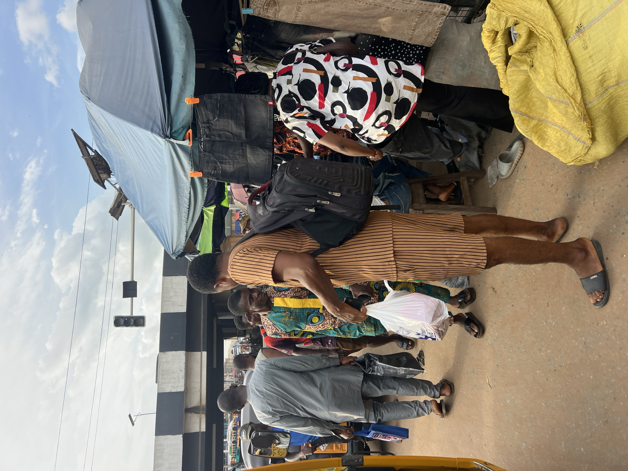 This image captures a busy street scene in Nkpor, Nigeria. Several people are visible, dressed in everyday clothing. The scene appears to be a market or a busy commercial area, with stalls or vendors set up along the street, indicated by the clothing racks and tarps. One person is wearing a patterned shirt and another is in a striped outfit, while others are in more subdued casual wear. A black backpack is slung over someone's shoulders. A yellow woven sack is prominent on the right side of the image. The ground is a dusty, earthy color. In the background, a traffic light is mounted on a pole, and a striped pedestrian bridge or overpass is visible. The sky is cloudy, suggesting it might be daytime. The overall impression is one of a bustling, active marketplace.