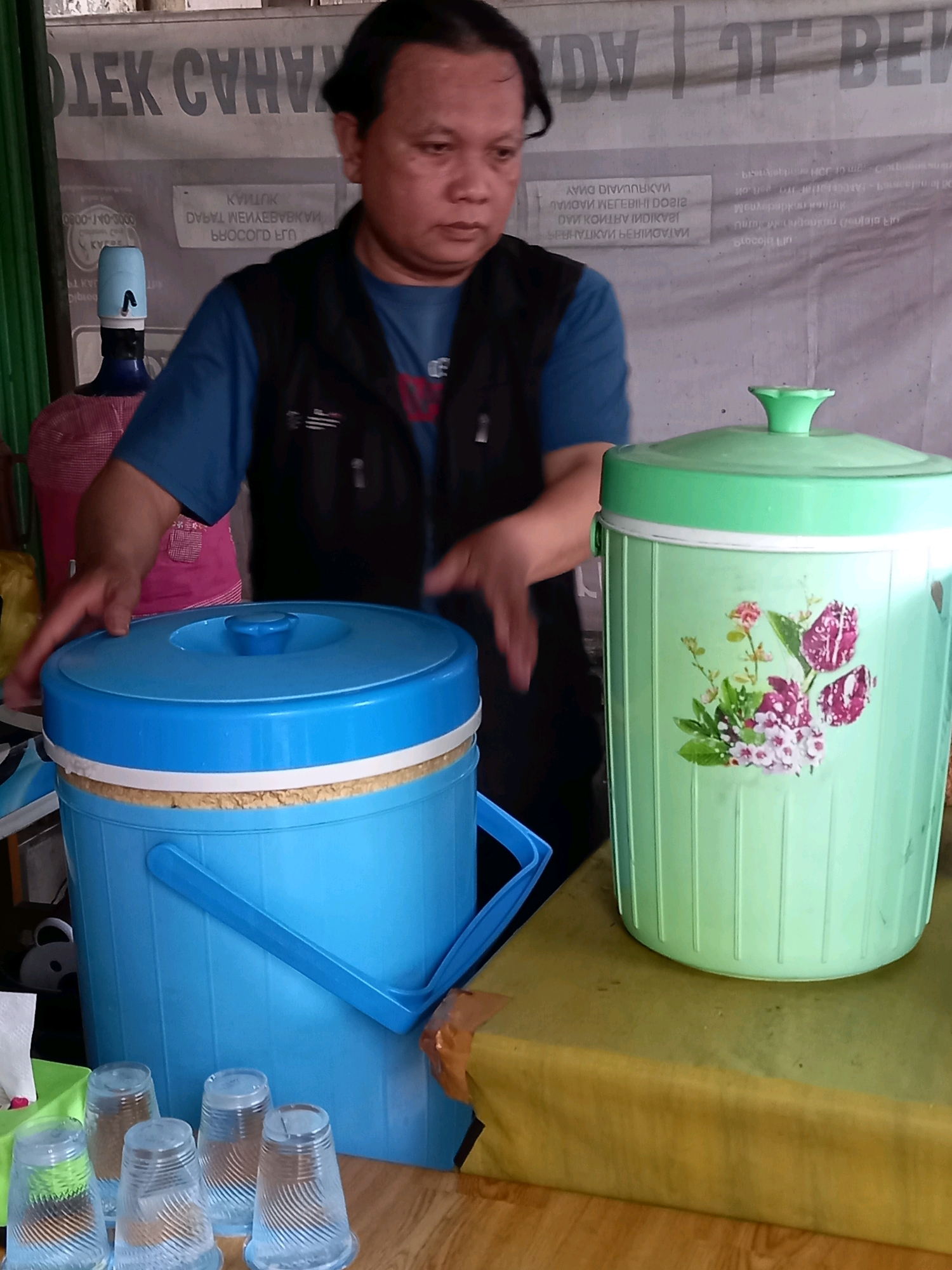 The image depicts an outdoor stall in Samarinda, Indonesia, likely a food or beverage vendor. A man with dark hair and a light complexion, wearing a blue t-shirt and a black vest, stands behind two large beverage dispensers. To his left is a light blue dispenser with a white rim, and to his right is a pale green dispenser adorned with floral decals. In the foreground, several clear plastic cups are neatly arranged on a table. The background features a banner with text in a foreign language, suggesting a commercial establishment. The overall setting appears to be a street or market area, typical of urban scenes in Indonesian cities.