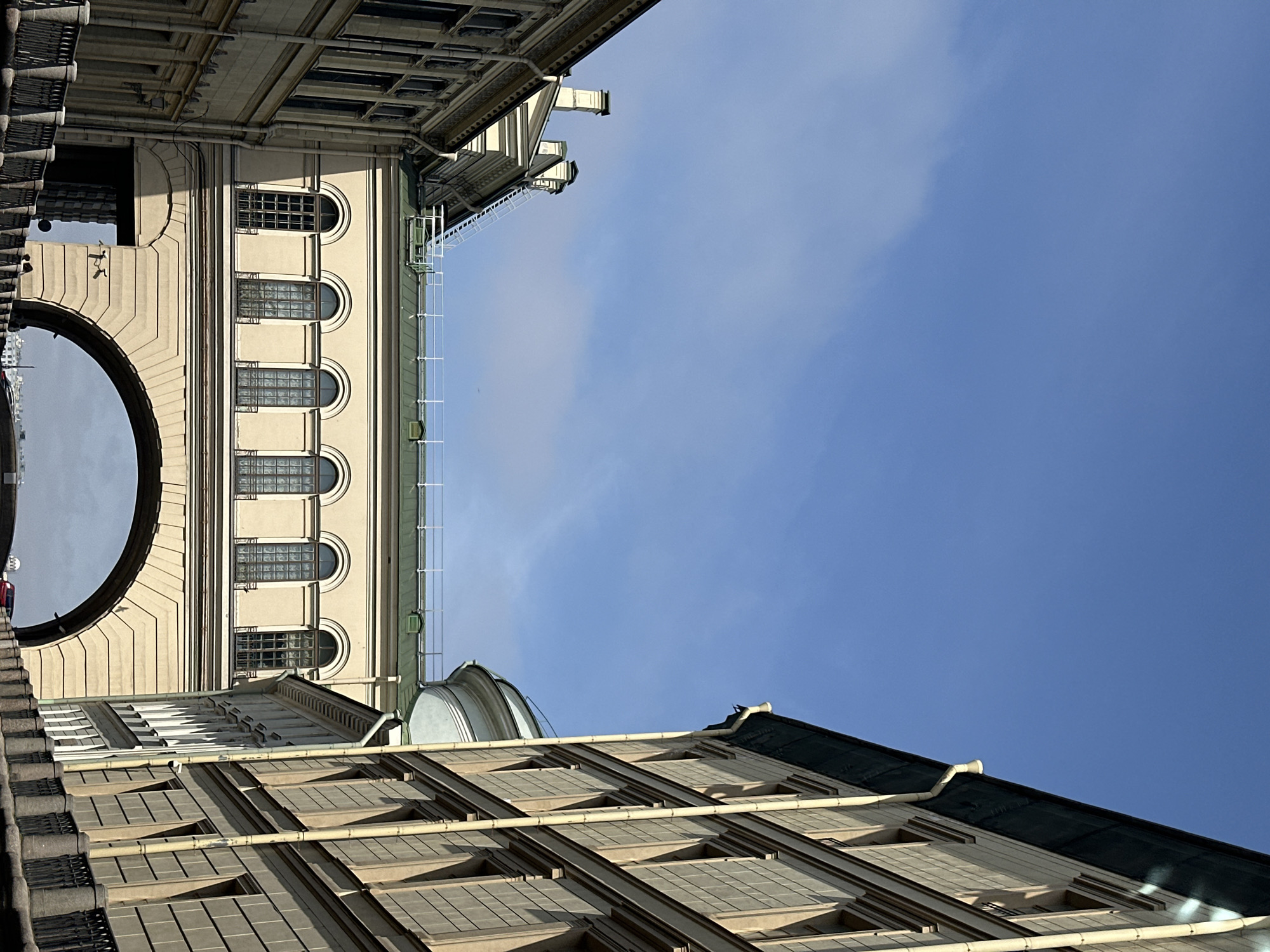 The image captures an upward-looking perspective between buildings in Saint Petersburg, Russia, dominated by a blue sky with a few scattered clouds. The buildings are constructed from a light-colored stone or concrete, featuring architectural details such as rows of arched windows, a dome, and a large arched opening. The buildings have a classical style, with repeating patterns of windows and cornices. The sky occupies a significant portion of the frame, emphasizing the height of the buildings and the open space between them.
