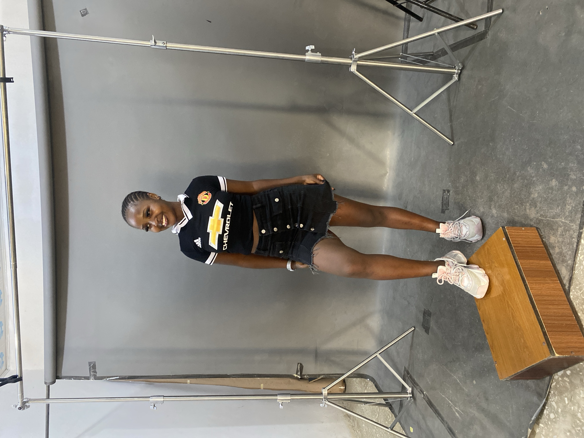A young Black woman stands in a photography studio. She is wearing a black Manchester United soccer jersey with white and yellow accents, a black denim mini-skirt with a distressed hem, and light pink athletic shoes. Her hair is braided in a cornrow style. She is smiling and looking to her left. She stands on a dark grey floor, with a grey backdrop behind her. To her left and right are professional photography light stands. In the foreground, to her left, is a wooden block. The location is Gura Top, Nigeria.