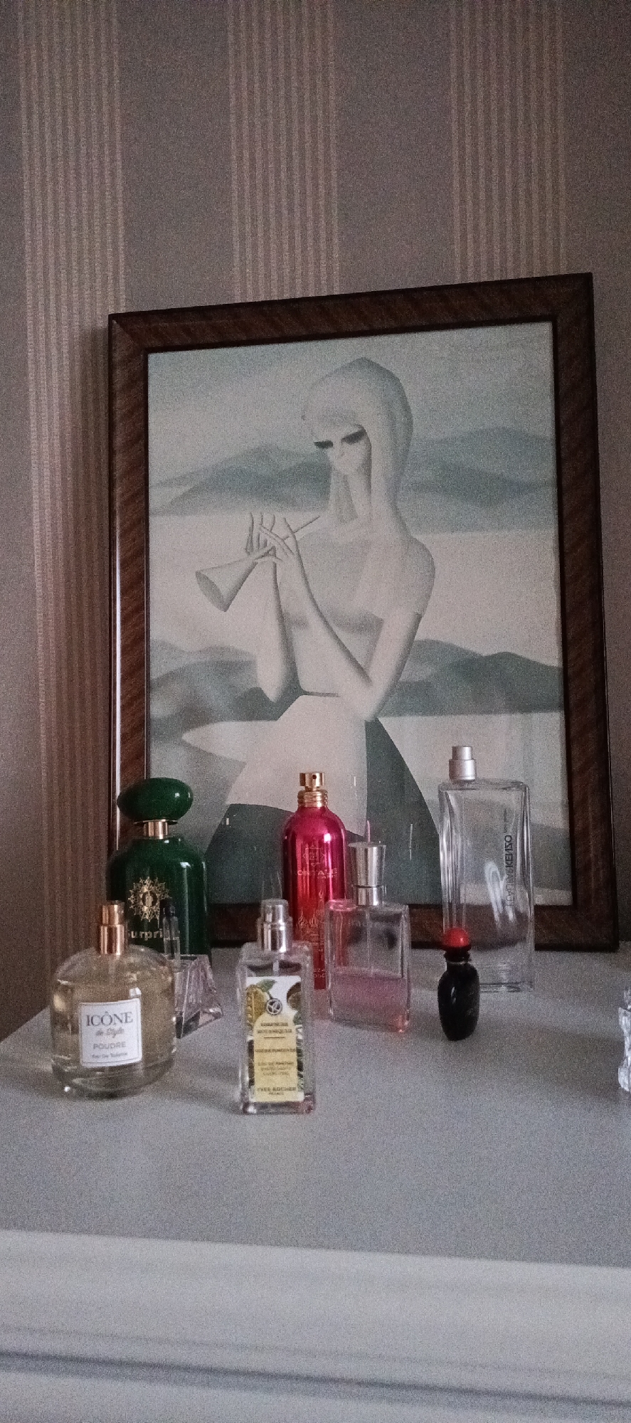 This image shows a collection of perfume bottles arranged on a shelf in front of a framed artwork. The artwork depicts a stylized figure in a veiled headdress playing a flute against a muted, mountainous background. The perfume bottles vary in size, shape, and color.
From left to right, the bottles are:
1. A large, emerald-green bottle with a rounded cap and a gold label.
2. A clear, round bottle with a gold cap, labeled "ICONE de LIGNE POUDRE Eau de Toilette".
3. A tall, rectangular bottle with a yellow label depicting botanical illustrations and text.
4. A bright pink, metallic bottle with a gold cap, labeled "ORIFLAME".
5. A clear, rectangular bottle containing pink-hued liquid and a silver cap.
6. A tall, clear rectangular bottle with a silver cap, labeled "KENZO".
7. A small, dark bottle with a red cap.
The setting appears to be indoors, likely a bedroom or dressing area, with striped wallpaper in the background. The lighting suggests it could be daytime, but the exact time is not determinable. There are no explicit cues about the weather or a specific location within Moscow, Russia, beyond the prompt's context. The arrangement of the items suggests a personal collection being displayed.