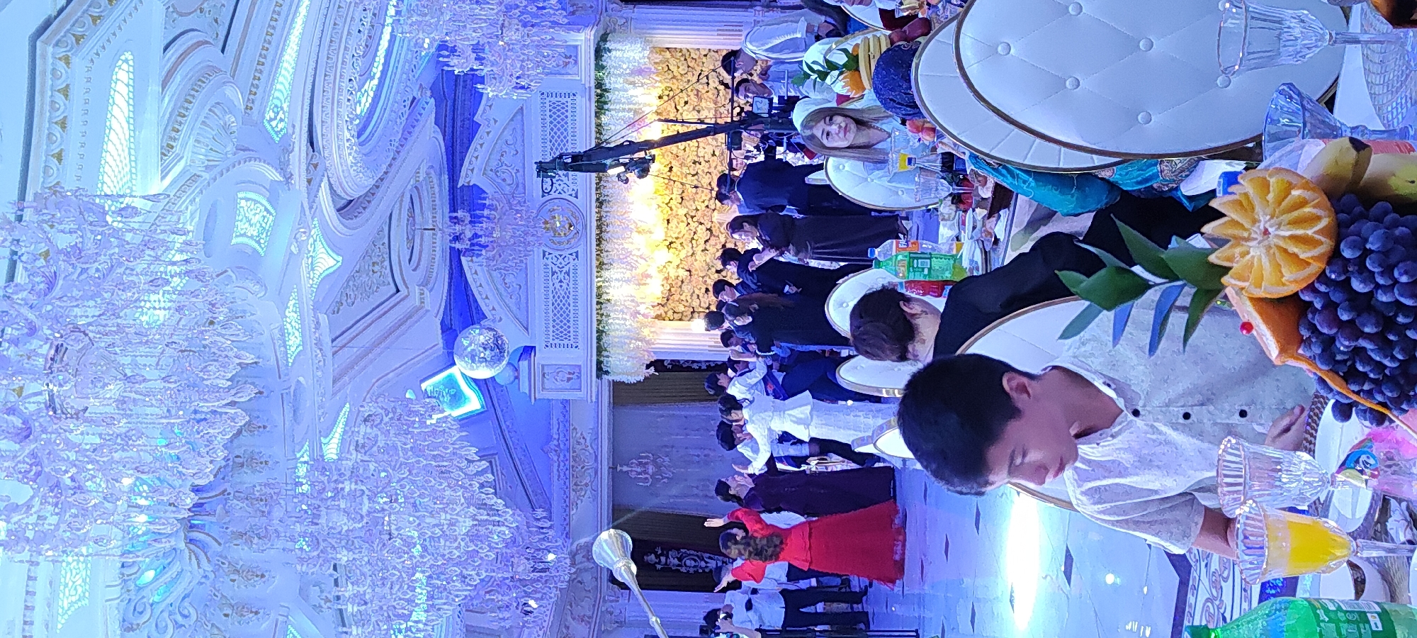The image depicts an indoor event, likely a wedding reception or formal celebration, taking place in a grand hall. The hall features an elaborately decorated ceiling with ornate patterns and multiple crystal chandeliers, casting a bright, blueish light over the scene. In the background, a large floral arrangement adorns the wall, illuminated by warm lights. A significant number of people are present, with some actively dancing in the center of the room, dressed in formal attire. Others are seated at tables, suggesting a dining or reception area. The foreground shows a young boy seated at a table, looking to his right. A fruit arrangement, including grapes, oranges, and a banana, is prominently displayed on the table in front of him, along with glasses and bottles of drinks. The overall ambiance appears festive and elegant.