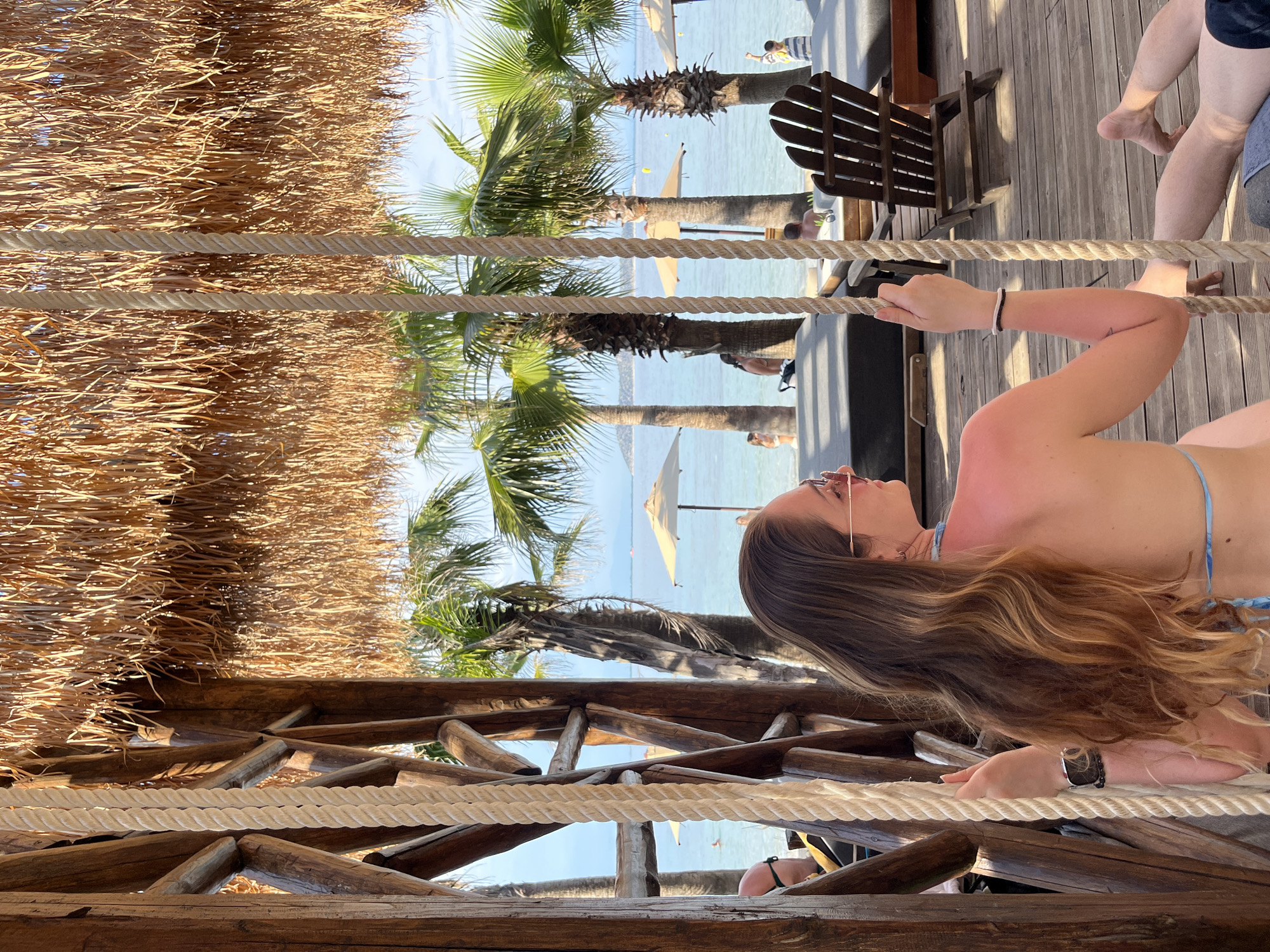 The image depicts a woman sitting on a swing, holding onto the ropes, in a beachfront setting. She has long, wavy brown hair and is wearing sunglasses. The swing is suspended from a thatched roof structure, with wooden beams and posts supporting the roof. In the background, palm trees line a beach with lounge chairs and umbrellas. The sea is visible, with a few people swimming in the distance. The overall atmosphere is relaxed and summery. The location is specified as the city of Thassos, Greece, which is an island in the northern Aegean Sea known for its beaches and natural beauty.