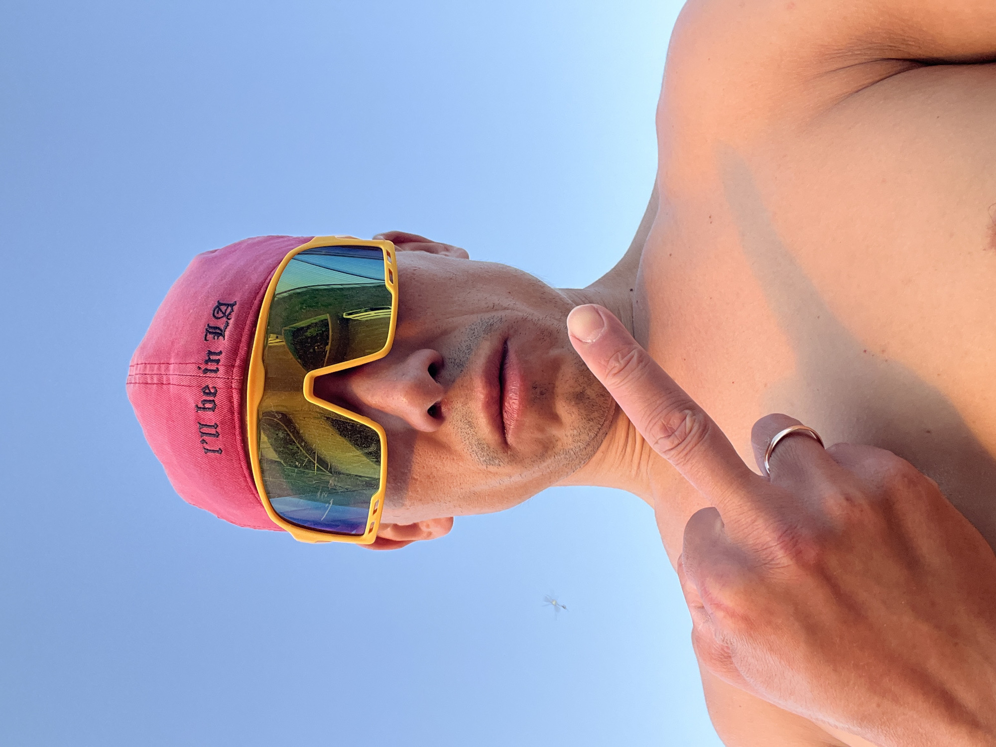 A man is posing for a low-angle selfie outdoors with a clear blue sky in the background. He is wearing a pink baseball cap with the text "I'll be in LA" embroidered on the left side and yellow sunglasses with mirrored lenses. He is shirtless and holding up his right index finger, which has a gold ring on it. An insect is visible in the background. The photo was most likely not taken in the city of Chaikovsky, Russia.
