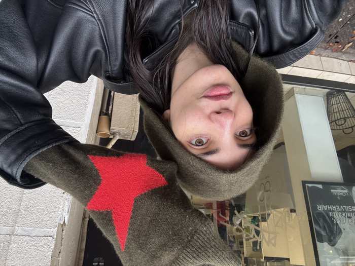 A person with dark hair and brown eyes is captured in a close-up, likely a selfie, on a street in Paris, France. They wear a dark green hooded garment and a black leather jacket. Their left hand is raised to head height, revealing an olive-green knitted glove adorned with a prominent red five-pointed star. The person exhibits a composed expression with slightly pursed lips.
In the immediate background to the left, a glass storefront is visible, displaying reflections of an interior. Text on the window reads "les fêtes, tombez du #SILVERHAIR" and includes the "L'ORÉAL" logo and a distinct "eB" logo, identifying the establishment as a hair salon. A black wire wastebasket sits near the bottom of the window. To the right, a textured, light-colored building wall is visible, with a small, light-brown cylindrical object on a ledge. The tiled sidewalk below shows some debris and a metal grate. The hair salon in the background is identifiable as eB Hair Coiffeur Visagiste, located at 10 Rue Jean Bologne, 75016 Paris.