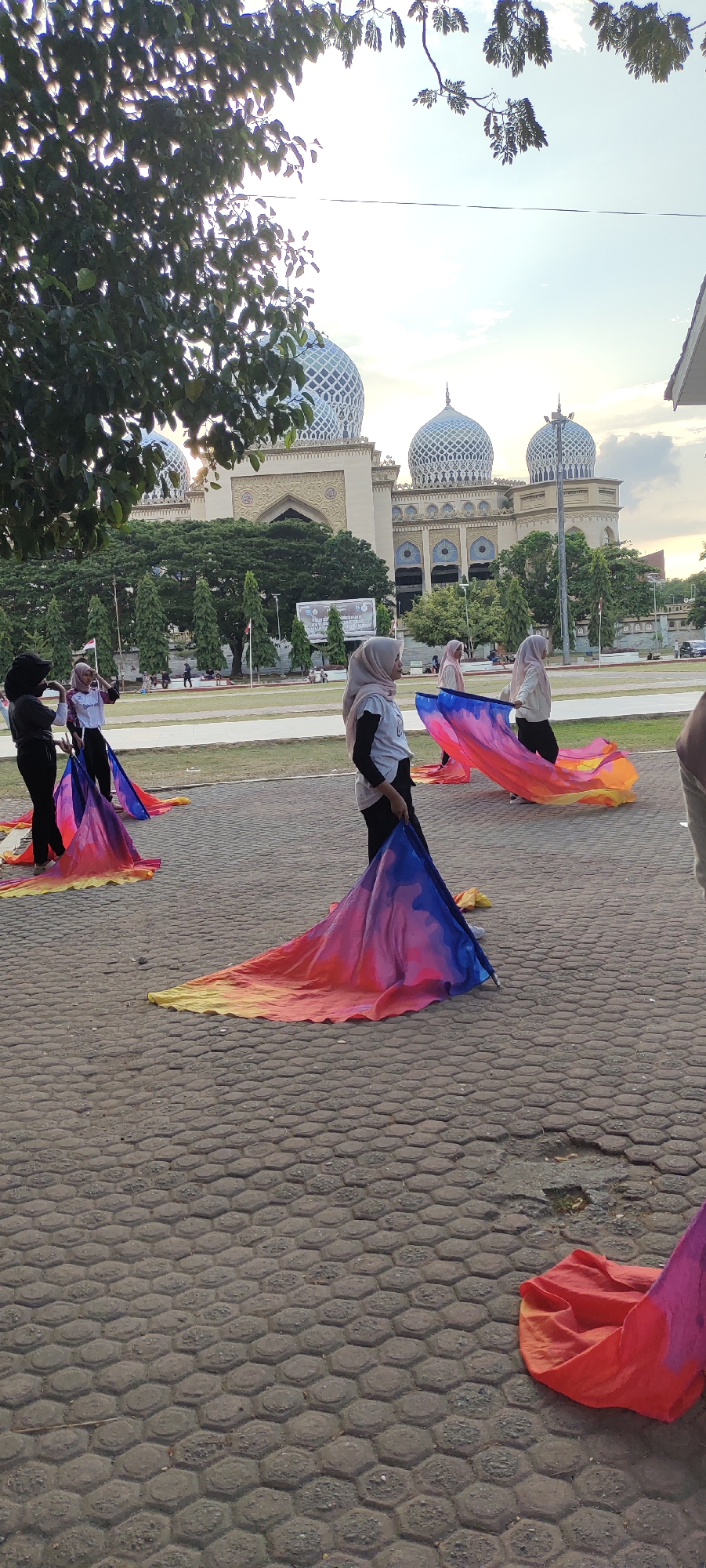 In Lhokseumawe, Indonesia, near the Baiturrahman Mosque, a group of women in hijabs practices with colorful flags. The flags, predominantly in shades of blue, pink, red, and yellow, are spread out on a hexagonal-patterned paved area. Some women hold the flags, which drape elegantly on the ground, suggesting a performance or rehearsal. In the background, the distinctive domes and architecture of the Baiturrahman Mosque are visible, along with trees and other people in the distance. The sky is partly cloudy, with the sun casting a warm light.