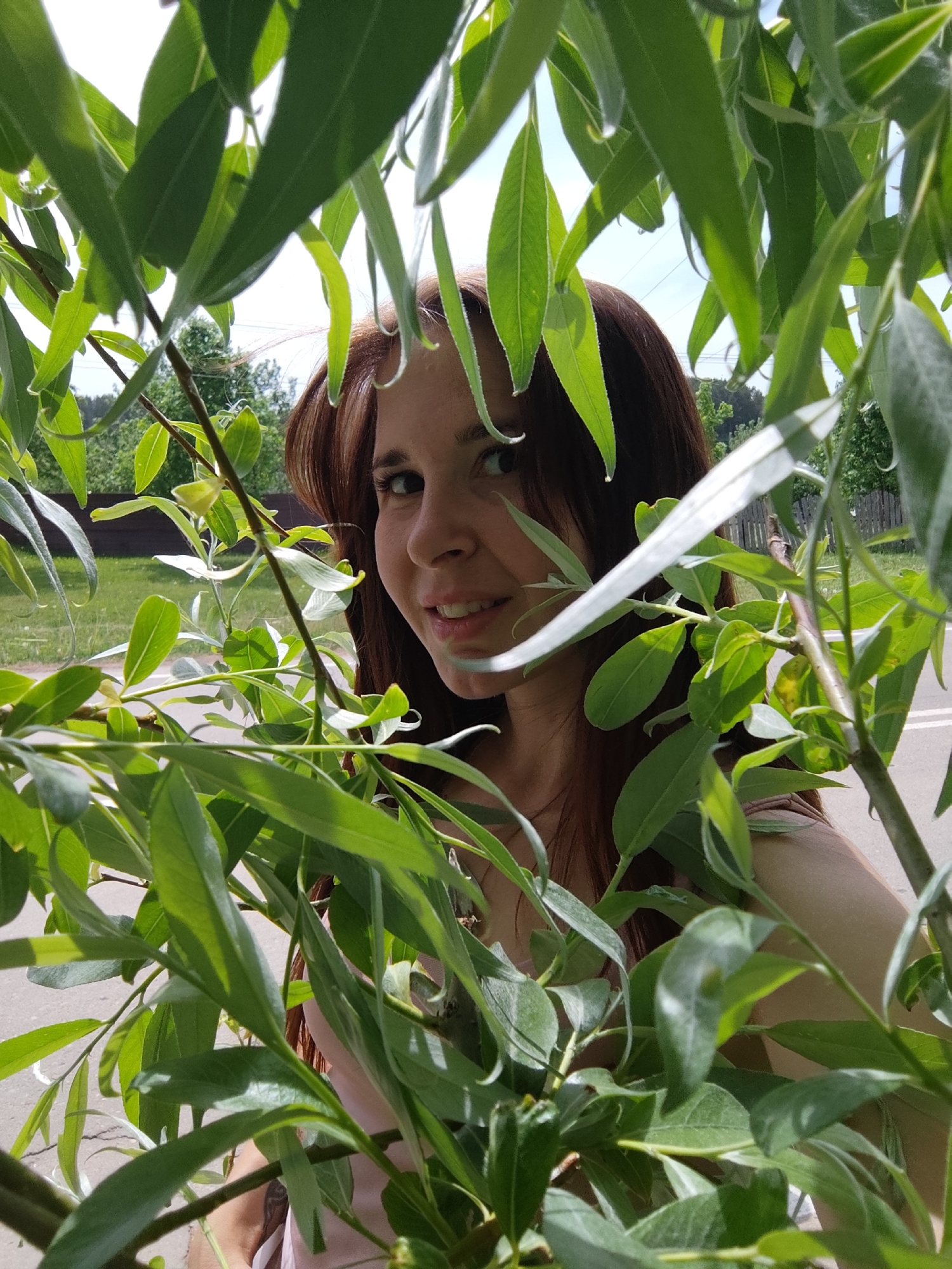 A young woman with dark hair and fair skin is partially obscured by the foliage of a tree, likely in a park or green area in Фоки, Russia. The woman smiles slightly toward the camera. Green leaves of the tree are in the foreground, both partially obscuring and framing her face. In the background, there is a road, a low fence, and more trees. The day appears sunny.