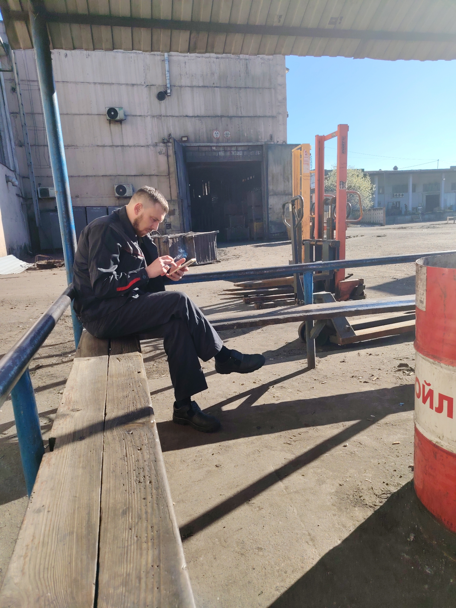 A man in a black jumpsuit sits on a wooden bench under a metal canopy structure. He is holding a mobile phone in his hands and appears to be looking at it. He is smoking. He has short hair and a beard. Behind him is a large gray industrial building with a large open doorway and two air conditioning units on the exterior wall. To the right of the man is an orange forklift and a red and white barrel with Cyrillic script on it. The ground is concrete and there are cigarette butts scattered around. The location is Noviy, Russia.