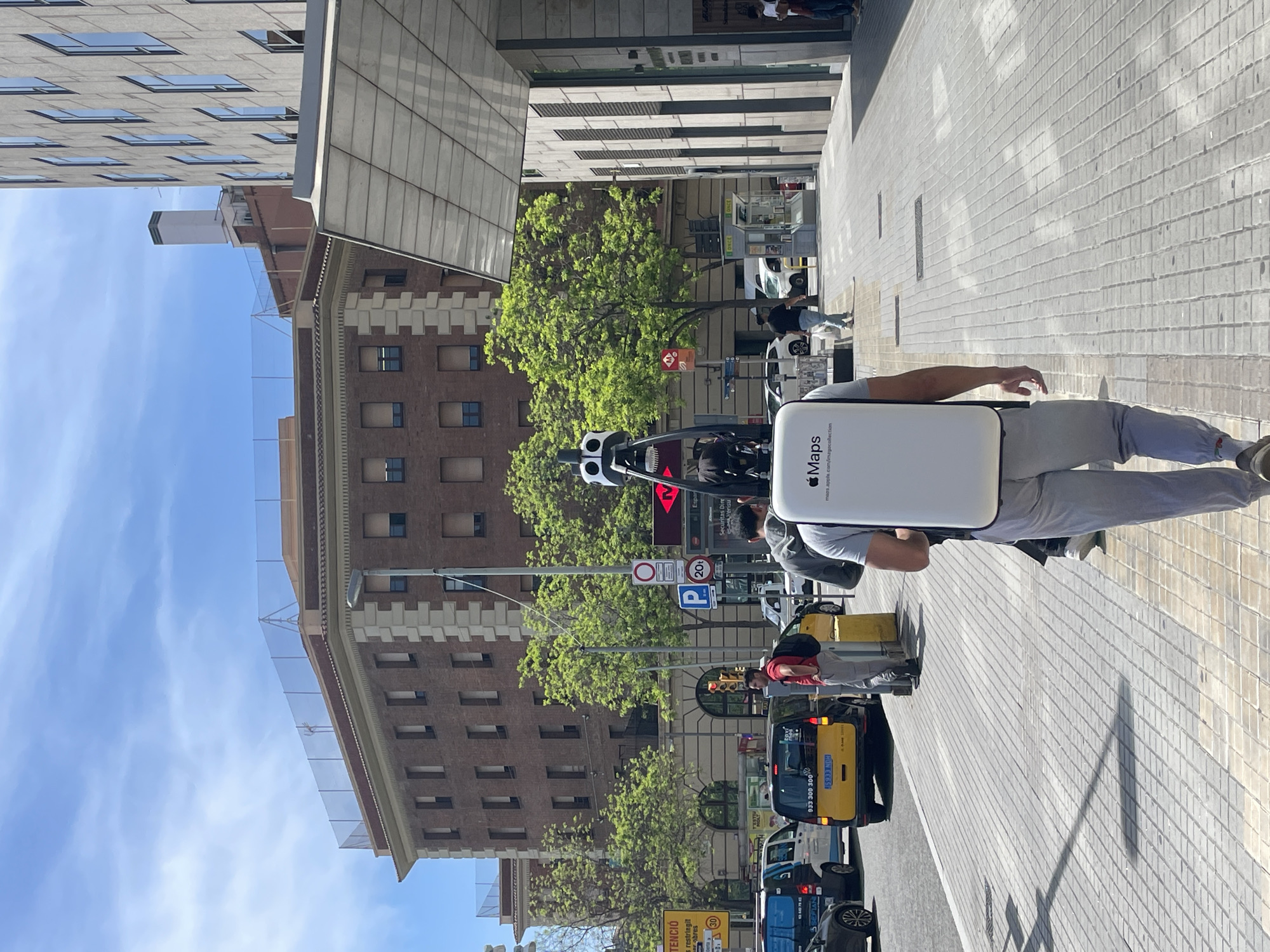 An upright, sunlit outdoor shot captures an urban street scene, likely in Barcelona, Spain. The primary subject is a person facing away from the camera, wearing a white backpack labeled "Maps" with "maps.apple.com/imagecollection" visible, and a helmet. This individual appears to be involved in street mapping, possibly riding a scooter or bicycle (partially visible).
The setting includes a wide pedestrian sidewalk paved with light-colored bricks on the right, and a street with vehicles on the left. Shadows cast by buildings and objects indicate bright midday sun. On the left side of the street, there's a modern building with many windows, and further back, a distinct older brick building with intricate architectural details and several windows. Lush green trees line parts of the street.
Several other people are visible: pedestrians on the sidewalk and a person in a red shirt near a yellow taxi. Other vehicles include a yellow taxi with text "933 300 300" and license plate "5933 NDH", and a blue vehicle with "SEFLAN".
Visible text and signs include "Maps" and "maps.apple.com/imagecollection" on the backpack. Street signs show a blue "P" for parking, "20" (possibly related to parking fees), and a red circular "no entry" sign. A yellow sign at the bottom left reads "ATENCIÓ trànsit restringit obres" (Attention, restricted traffic works) and "30" (likely a speed limit). Text like "ONCE", "Securitas Dire", and "Espan" also appear on kiosks or buildings. The clear blue sky indicates fair weather.