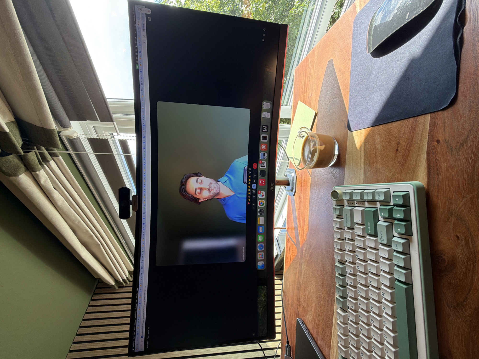 A man is on a video call on a curved monitor. The monitor displays the man, who is wearing a blue shirt. The monitor is on a wooden desk. Also on the desk is a keyboard with green and white keys, a mouse, a mousepad, and a glass of coffee. The scene appears to be in an office or home office, with blinds and a window in the background.