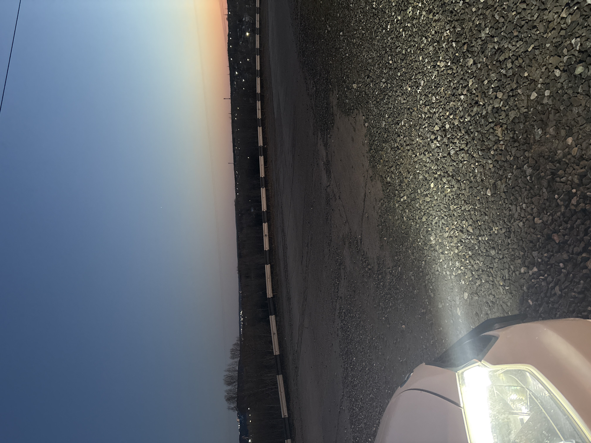 Here is a detailed description of the image:
**Scene Overview:**
The image appears to be taken at dusk in a paved or partially paved area with gravel present. The horizon shows a color gradient from orange to blue, indicating the time of day. A car with its headlights on is visible in the foreground.
**Location:**
While the image itself doesn't provide definitive location data, the prompt mentions Chaikovsky, Russia. Without further details within the image, it's impossible to pinpoint an exact location in the city. The scene suggests an area possibly on the edge of town, near a road or parking area.
**Subjects and Objects:**
* **Car:** The front part of a car is visible in the lower right of the image. Its headlights are turned on and illuminating the ground.
* **Headlights:** The bright light from the headlights shines onto the gravel surface, providing a focused area of illumination.
* **Gravel:** A large portion of the image shows gravel surface, possibly a parking lot, driveway, or roadside area.
* **Pavement:** A paved surface exists, with cracks visible.
* **Road Barrier:** A road barrier runs along the side of the road.
* **Horizon:** The horizon line is visible with a gradient of colors, signifying the sunset or dusk.
* **Distant Lights:** There are some distant lights on the horizon, likely from buildings or streetlights.
**Environment:**
* **Lighting:** The scene is dimly lit due to the time of day. The car's headlights provide the primary source of light in the foreground.
* **Weather:** The weather is not specifically indicated, but the clarity of the image suggests relatively clear conditions.
**Actions and Interactions:**
* **Car Operation:** The car's headlights are turned on, indicating operation or preparation for operation.
* **Light Emission:** The headlights emit a concentrated beam of light onto the gravel.
**Summary:**
The image depicts a car at dusk in an area with gravel. The car's headlights illuminate the scene. Without additional details, the precise location in Chaikovsky, Russia, cannot be determined.