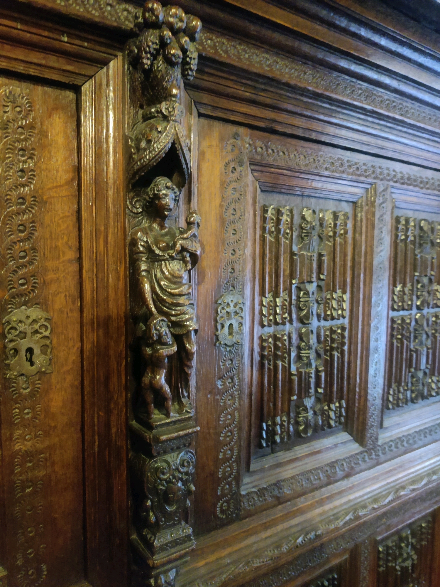 The image shows a close-up view of intricate dark wood carvings on a piece of furniture, likely a cabinet or armoire. The carving features figures, scrollwork, and decorative patterns. On the left, a wooden carving of a woman holding a child is prominent, with another smaller figure at her feet. To the right, the furniture has panels with detailed, repetitive patterns that appear to be relief carvings with some gilded elements. The wood is polished and has a rich, aged appearance. The overall impression is one of detailed craftsmanship and historical design. This object is located in the Rijksmuseum in Amsterdam, Netherlands.