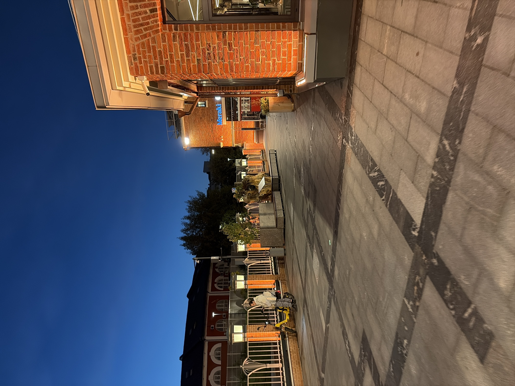 An outdoor shot captures a twilight scene in Moscow, Russia. The sky is a deep, clear blue. In the foreground, a paved walkway features a pattern of darker, inlaid lines. To the left, a person is paused on a yellow electric scooter near a decorative fence. Behind the fence, several ornate streetlights cast a warm glow. Further back, a building with red brickwork and arched windows is visible. To the right, another red brick building is illuminated by interior lights and features a prominent blue sign with the word "Meraki." The scene is tranquil, with no other people visible.