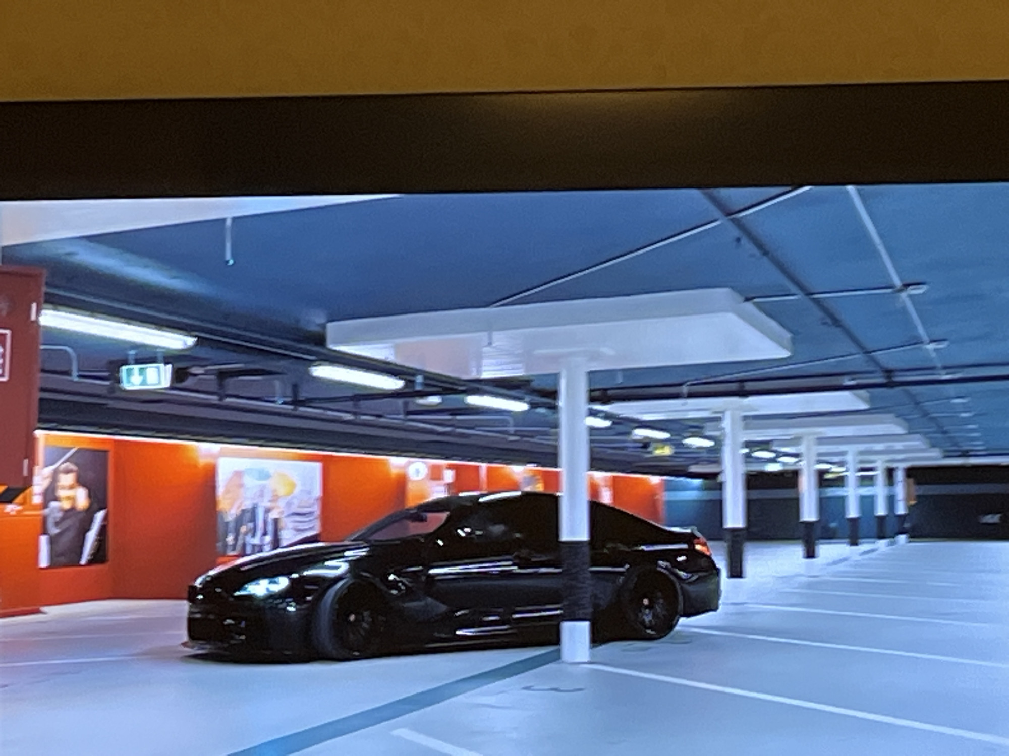 The image depicts a black car parked in what appears to be an indoor parking garage. The car is angled towards the viewer, with its front right tire turned slightly. The parking garage has a blue ceiling with fluorescent lighting and white pillars supporting the structure. The floor is marked with white parking lines. On the left side of the image, there are two framed pieces of art displayed on an orange wall, and a red structure with a white sign. The overall lighting suggests it is daytime, but the interior environment indicates controlled lighting conditions. There is no visible indication of the city of Kharkiv, Ukraine, or any specific geographical features that would confirm its location.