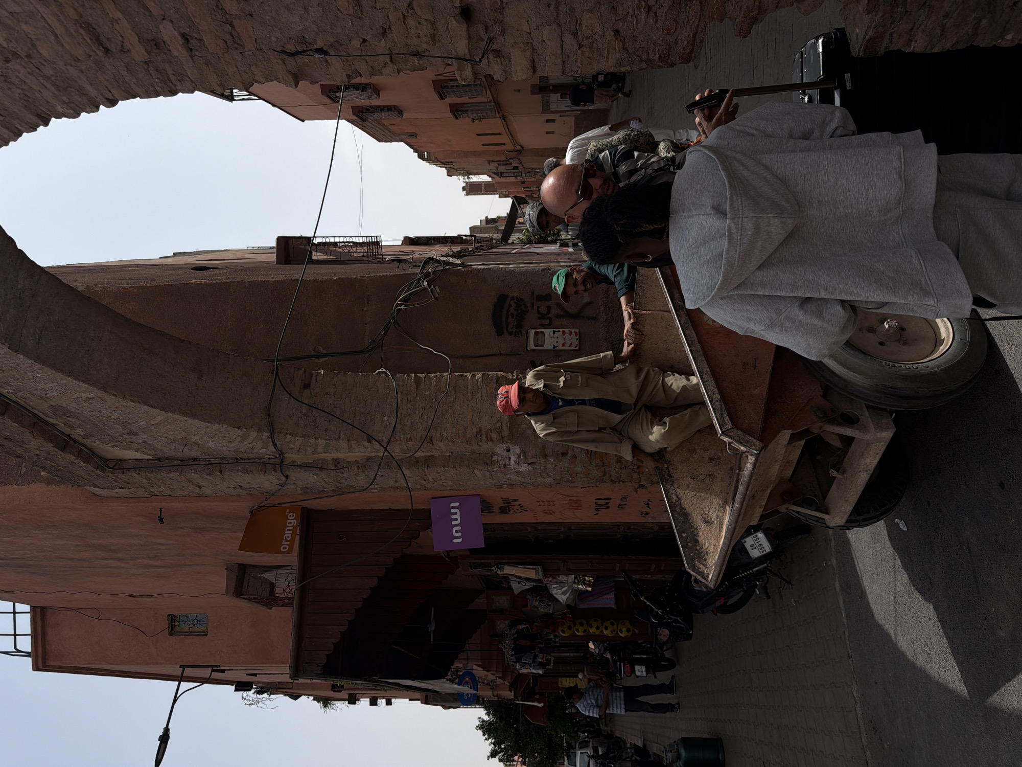 In Marrakesh, Morocco, a sunny day casts shadows on a narrow street lined with aged, terracotta-colored buildings and a large arched passageway. Several people are present: a man in a tan uniform and red cap sits in a rustic, orange-brown industrial cart, his legs visible; a younger person in a grey hoodie and sweatpants stands facing away from the camera, holding a dark suitcase on its wheels, with their left hand near the cart; a bald man wearing sunglasses and a striped shirt is looking towards the foreground; a man in a green cap is smiling; and another person in a white shirt stands behind them. Other individuals are visible further down the street. The environment includes shops with wooden overhangs, motorcycles parked on the side, and visible signage, including an "orange" logo and a purple "inwi" logo. The ground is paved with brick and asphalt, with tire tracks visible. The overall scene captures a moment in a bustling, historic urban setting.