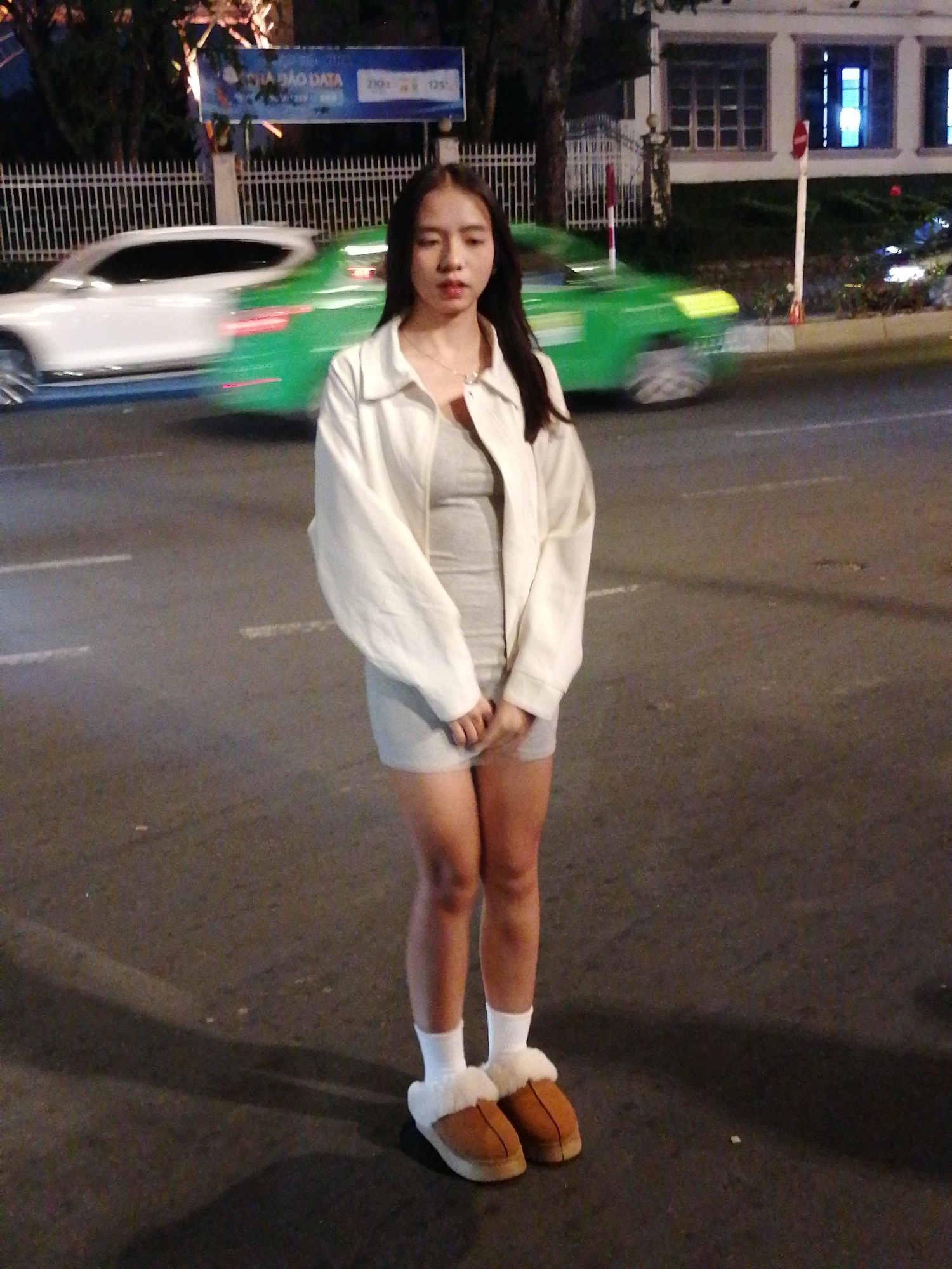 A full-body, medium-shot photograph depicts a young Vietnamese woman standing in the middle of a street at night in Da Lat, Vietnam. The woman has long, dark hair and is wearing a light-colored jacket over a grey dress and white socks with brown fuzzy slippers. She is looking directly at the camera with a neutral expression. In the background, there are two moving vehicles: a white car on the left and a green taxi on the right. The street is paved, and there are streetlights and buildings visible in the distance. A large digital billboard with Vietnamese text is also present. The overall scene suggests an urban environment at night.