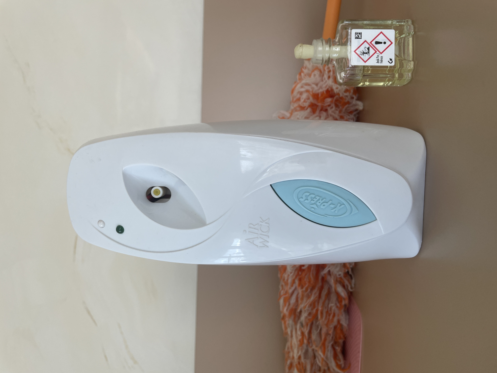 A close-up, horizontally oriented image captures various household items in an indoor setting. The primary focus is a white, oval-shaped automatic air freshener dispenser, likely mounted on a wall, positioned slightly left of center. The dispenser features a light blue button with "X-PRESS" embossed on it, along with the "AIR WICK" logo printed in grey lettering on its body. A nozzle with a yellow/green center is visible on the left side of the dispenser, and two small indicator lights, one clear/white and one green, are present below it.
To the right and slightly behind the air freshener, a clear, rectangular glass bottle containing a light yellow liquid is visible. This bottle is attached to a white/yellow dispenser mechanism and features a prominent label. The label displays several hazard pictograms: a GHS02 flame symbol indicating flammability and a GHS07 exclamation mark symbol indicating an irritant or harmful substance. Text "Ambii Per / fabrices" is also visible on the label, along with a QR code and a curved arrow. The bottle appears to be part of a plug-in or refill system.
Partially visible beneath and behind the bottle and air freshener is a fluffy cleaning tool, possibly a duster or mop head, characterized by its vibrant orange and white yarn-like fibers. The background consists of two walls: a light beige or off-white wall on the left, showing subtle, faint vertical streak patterns, and a solid light brown or darker beige wall on the right. The lighting is even and bright, suggesting an indoor environment. No people are present, and no specific activity is taking place; the image captures a still arrangement of these objects.