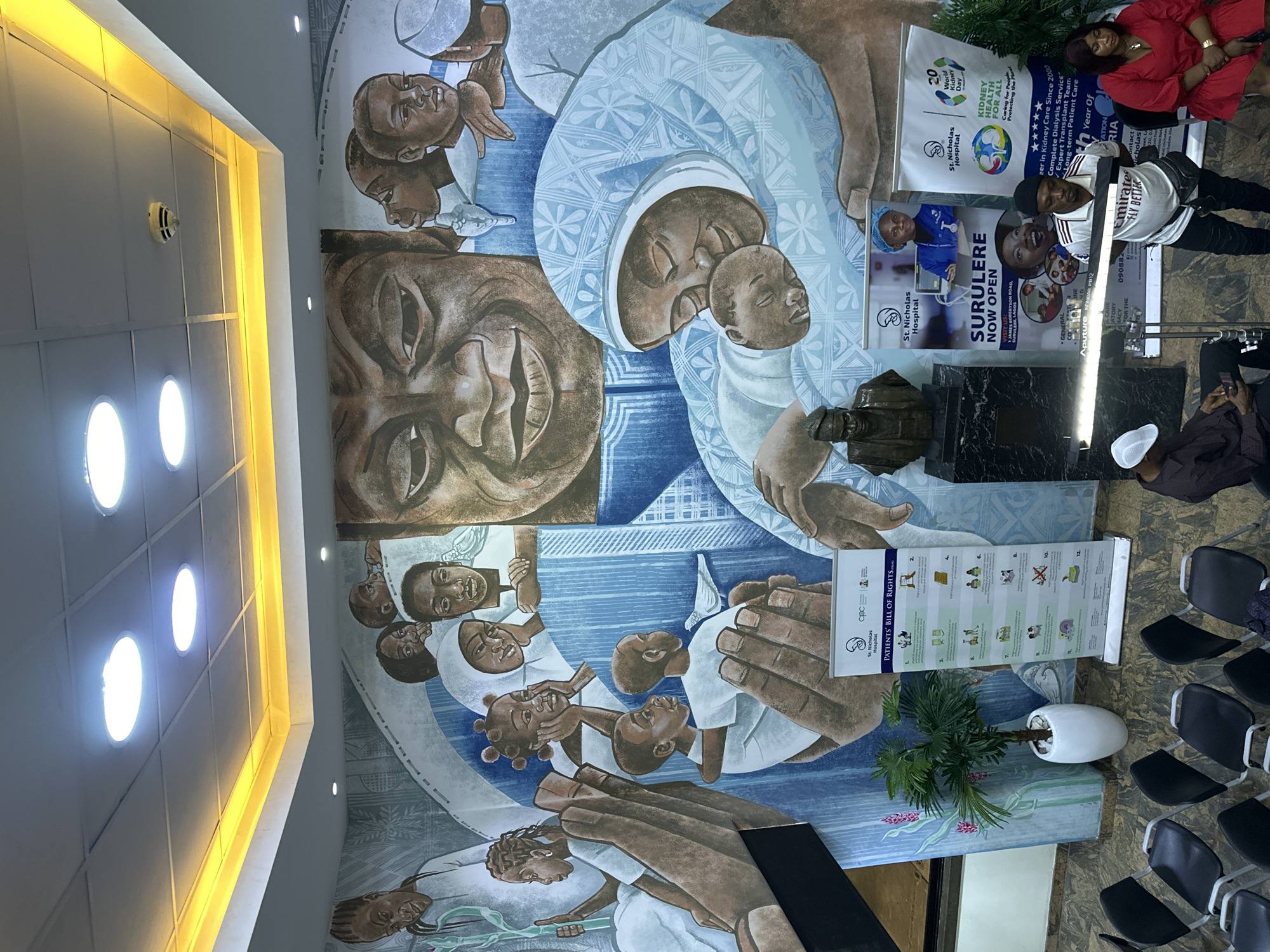 The image captures the interior of a St. Nicholas Hospital location, likely a waiting area, in Surulere, Lagos, Nigeria. A large mural depicting dark-skinned figures, some holding babies, dominates the wall. Displayed in front of the mural are posters for "World Kidney Day," information about a "Surulere Now Open" location, and a "Patients' Bill of Rights." A bronze bust is also present. Several people are present, including two women standing and posing for a photo and a man seated and using his phone. Rows of black chairs line the side of the room, indicating a waiting area. Overhead, the ceiling features square panels with embedded lights.