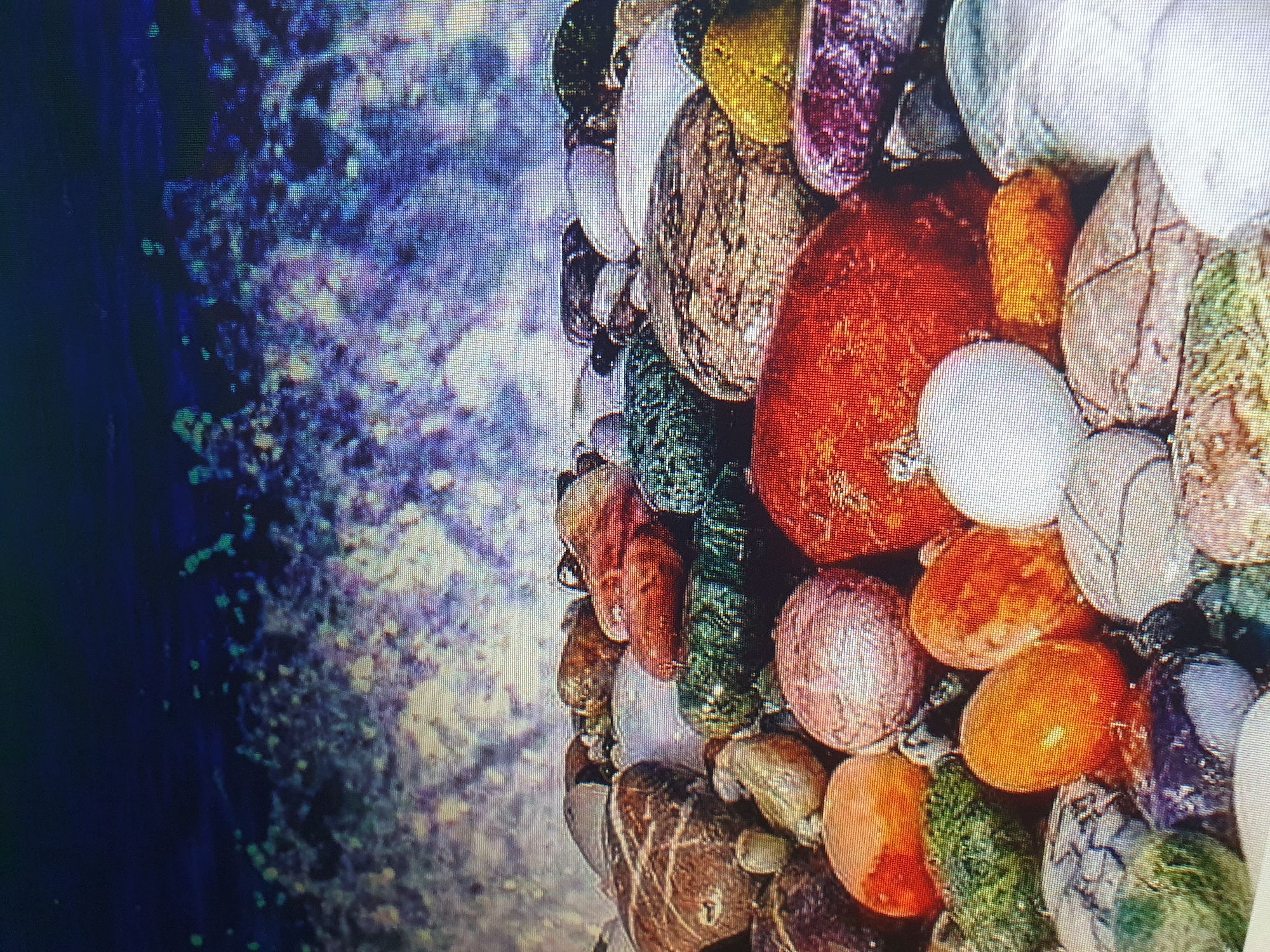 The image depicts a close-up view of colorful, smooth, rounded stones, likely from a riverbed or beach, against a blurred background of water and foam. The stones display a variety of colors including red, orange, green, purple, white, and gray, with some exhibiting intricate patterns. The foreground is densely packed with these stones, overlapping and creating a textured surface. The upper portion of the image shows the dynamic movement of water, with splashes and waves rendered in shades of blue and white, suggesting a natural environment. There are no people or man-made objects visible in the scene. The scene is presented in a natural, artistic style, focusing on the textures and colors of the stones and water. There is no information to suggest a specific location in Tashkent, Uzbekistan.
