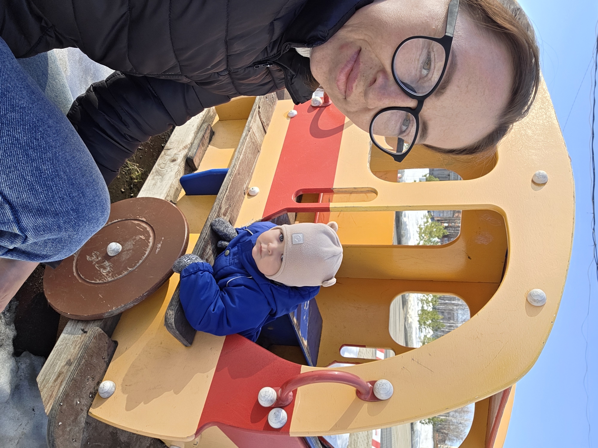 An outdoor scene in Chaikovsky, Russia, depicts two individuals near a children's play structure on a sunny day. In the foreground on the left, an adult female is partially visible, oriented towards the camera. She wears black-framed glasses, a black quilted jacket over a white collar, and blue denim jeans. Her dark hair is pulled back, and she presents a neutral expression.
To the right of the adult, a young child, appearing to be an infant or toddler, is seated inside a yellow and red play structure. The child wears a blue hooded snowsuit, a light tan knit beanie with small ear-like protrusions, and grey patterned mittens. The child looks towards the left with a solemn expression.
The play structure, likely made of wood or plastic, is predominantly yellow with red accent panels and features that resemble a small vehicle or train car. It includes a visible brown wheel on its lower right side and a red grab handle. The ground surrounding the structure consists of patches of melting snow, ice, and exposed dark earth. In the background, a paved area or road is visible, flanked by scattered trees, some coniferous and others deciduous with bare branches. Distant buildings are discernible under a clear blue sky.