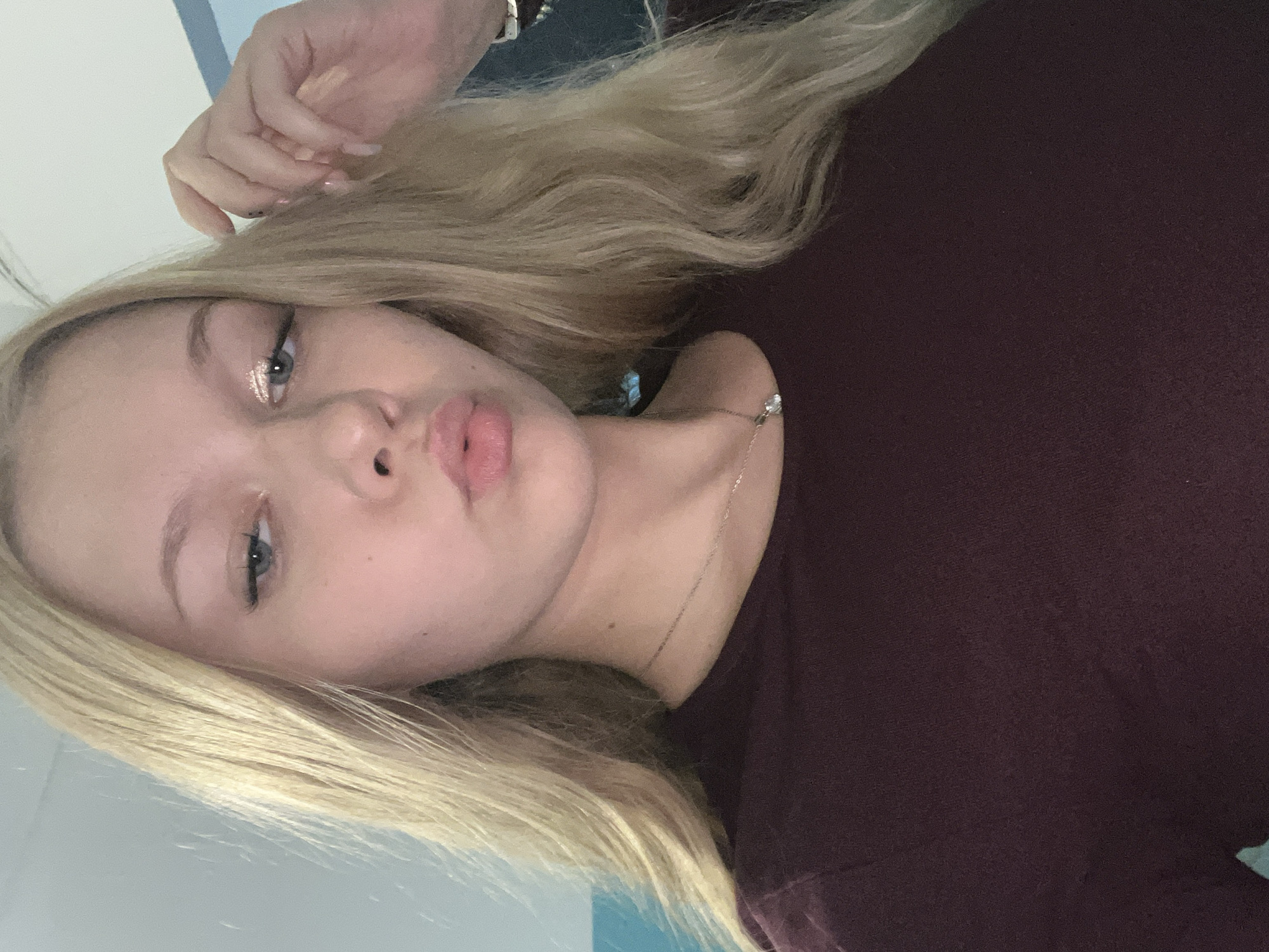 A young woman with long blonde hair and fair skin is captured in a close-up, selfie-style photograph. She is wearing a dark red, long-sleeved shirt and a delicate silver necklace with a pendant. Her makeup includes subtle eyeshadow and mascara. She is looking directly at the camera with her lips slightly puckered. Her right hand is raised, touching her hair. The background is a soft, light blue wall. There is no discernible location within the city of Perm, Russia, or any other specific geographical context evident in the image.