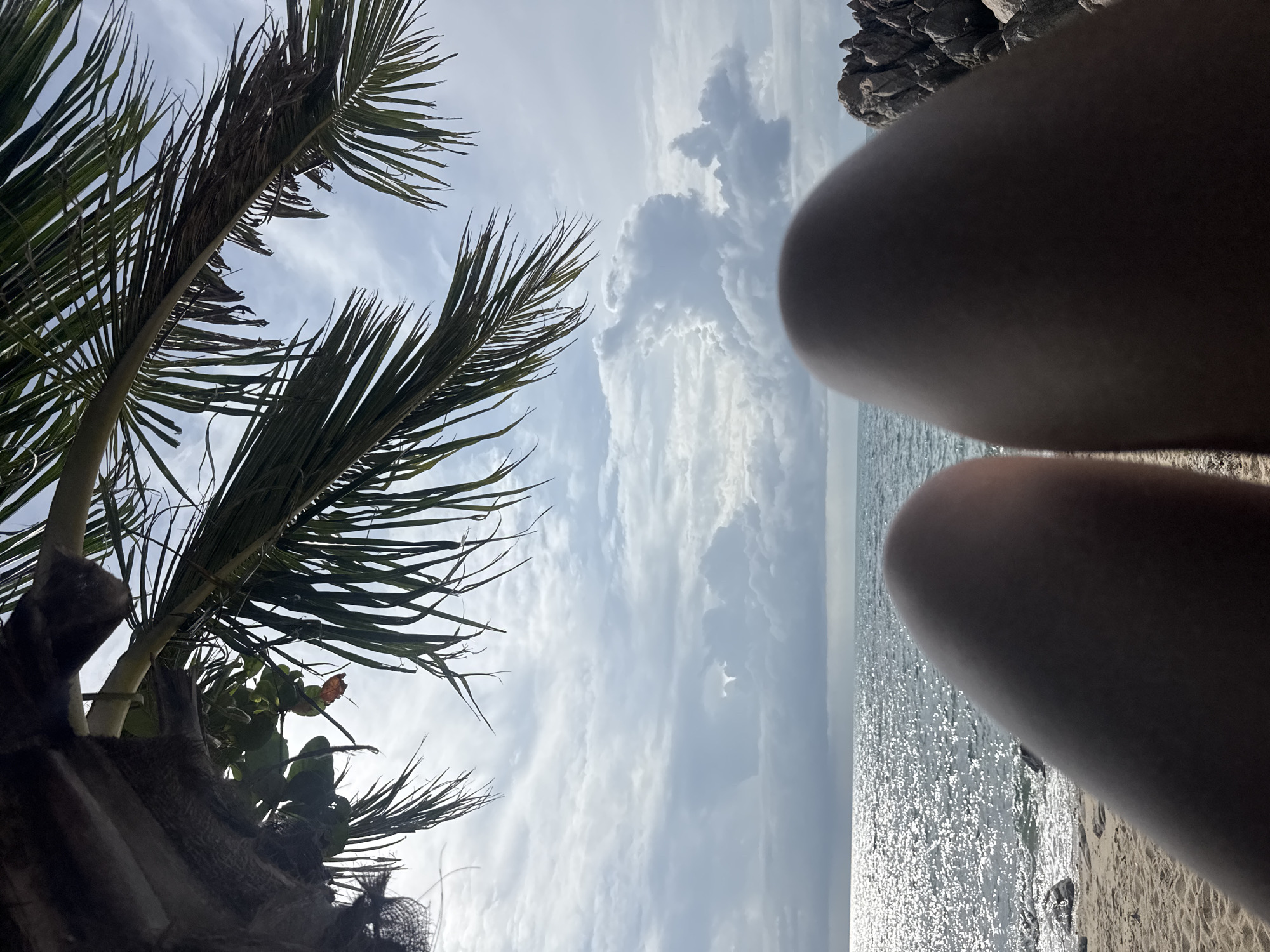 A first-person perspective shot shows the legs of a person on a sandy beach in Ko Pha-ngan District, Thailand. The legs are positioned in the foreground, obscuring the view of what the person is looking at. Beyond the legs, the scene opens up to a calm ocean with shimmering sunlight reflecting off the surface. A few rocks are visible on the right side of the beach. Overhead, palm fronds from a nearby tree frame the sky, which is filled with dramatic clouds. The overall impression is one of relaxation and enjoying a tropical beach setting.