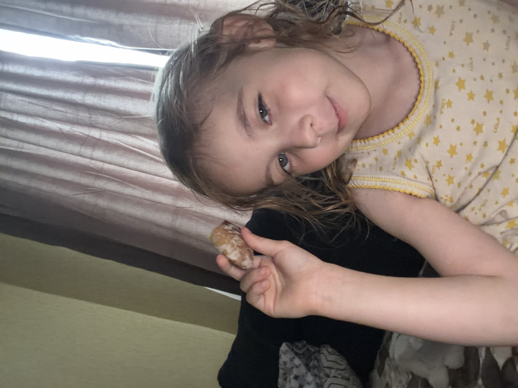 A young girl with light brown hair, wearing a yellow tank top with a star pattern, holds a small piece of food in her right hand near her face. She is looking towards the camera with a slight smile. The background consists of beige and pink curtains and a dark surface that appears to be furniture. The image was taken indoors, likely in a residential setting. The location in Tambov, Russia, is not determinable from the image itself.