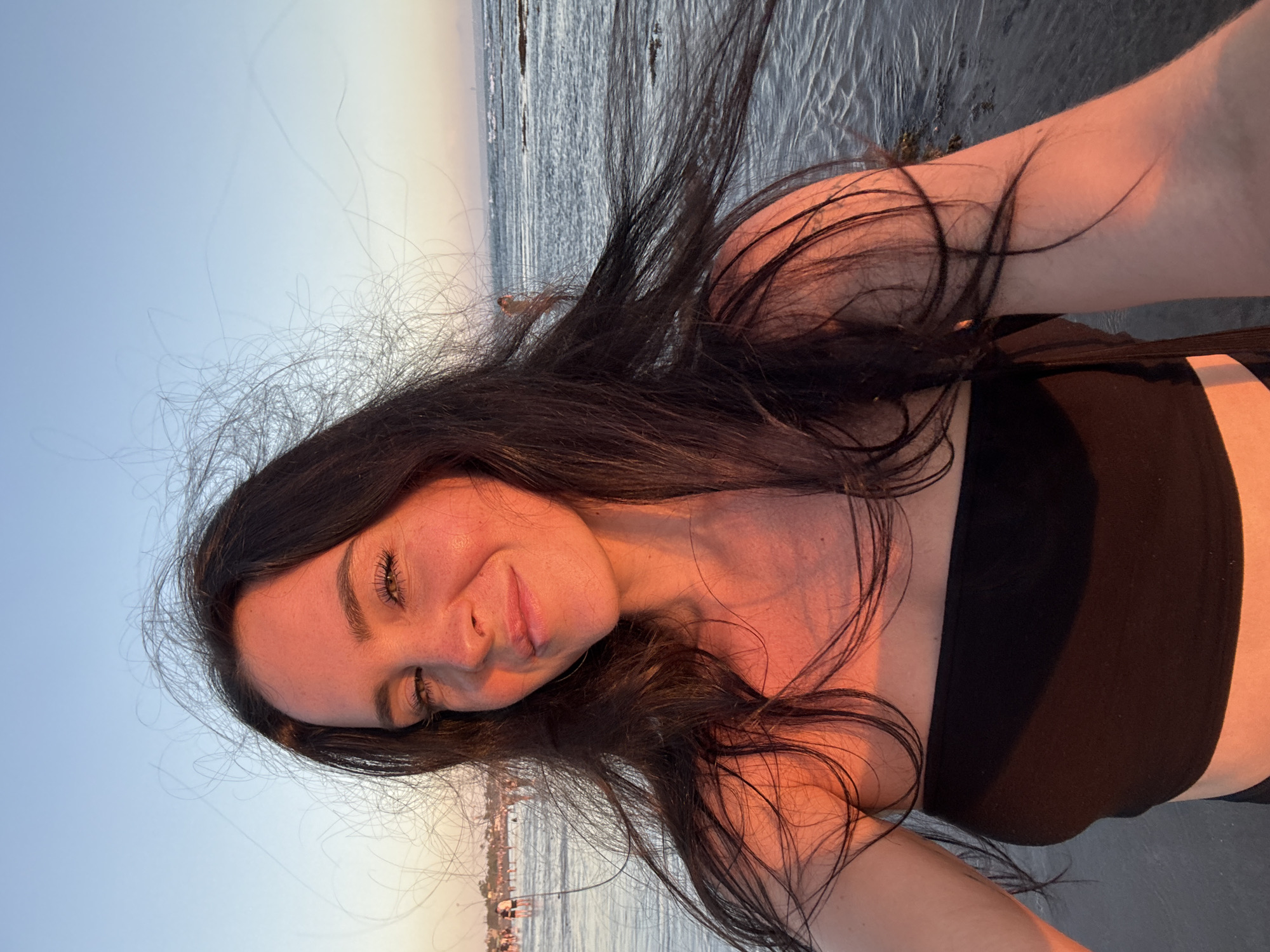 This is a close-up, vertical, selfie-style photograph of a young woman with long, dark, wavy hair. She is outdoors, with a body of water and a distant shoreline visible in the background. The sky is a pale blue, suggesting it is either sunrise or sunset, with a warm glow illuminating the woman's skin. Her eyes are open and she is smiling softly. She is wearing a black, strapless top. The hair is blowing slightly, which, along with the warm lighting, suggests a breezy environment. The scene appears to be a beach or coastal area. No visible text is present in the image. The overall mood is relaxed and serene, capturing a moment of quiet enjoyment in a natural setting.