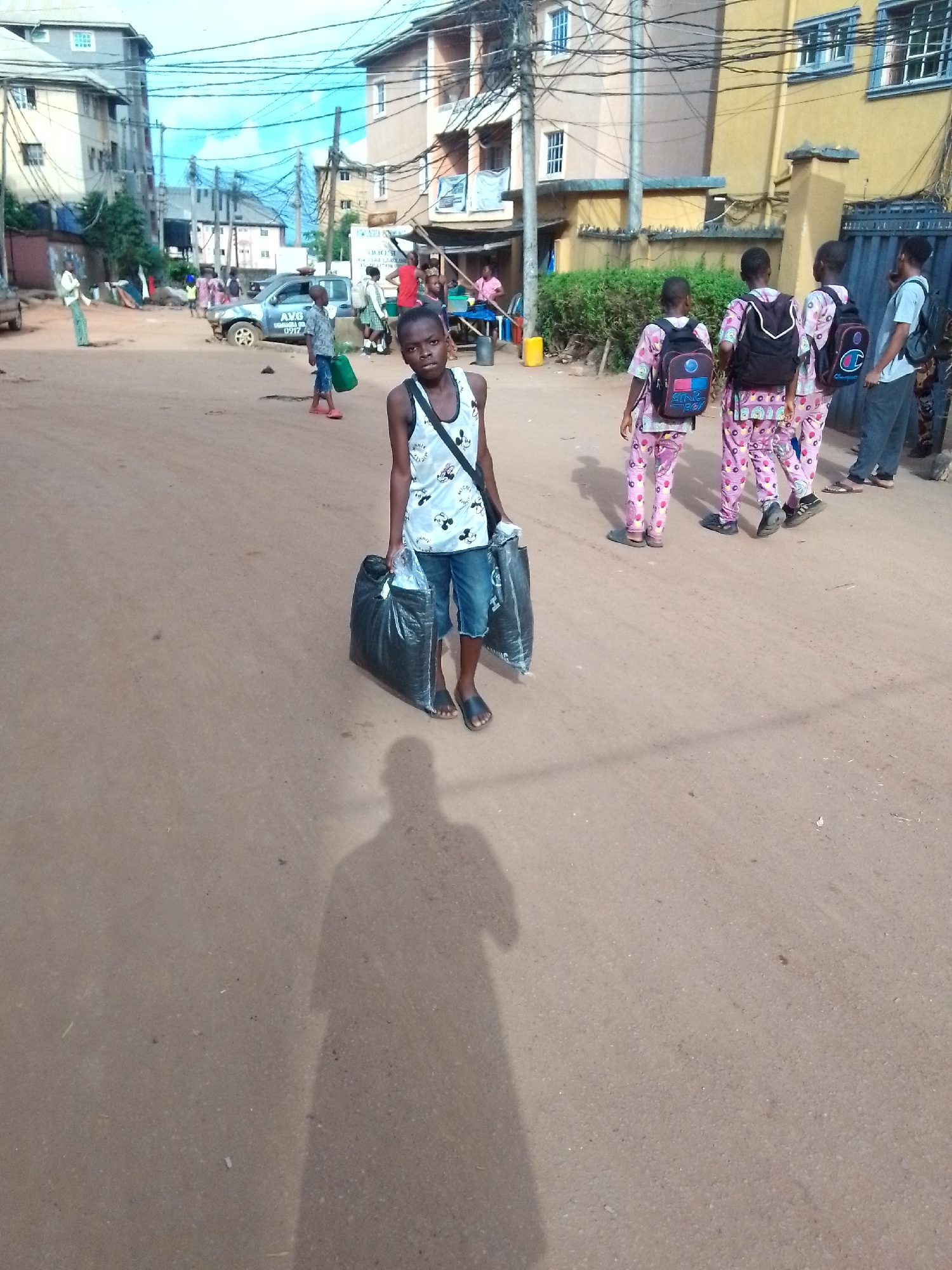 In a street in Onitsha, Nigeria, a child in a white Mickey Mouse tank top and denim shorts carries two large black plastic bags. His shadow is prominently cast on the dusty road. Nearby, three children in matching pink pajamas with backpacks walk with a man. Other people and vehicles are in the background, with buildings visible in the distance. Overhead power lines crisscross the scene. A sign advertising "AVA" is visible near a vehicle.