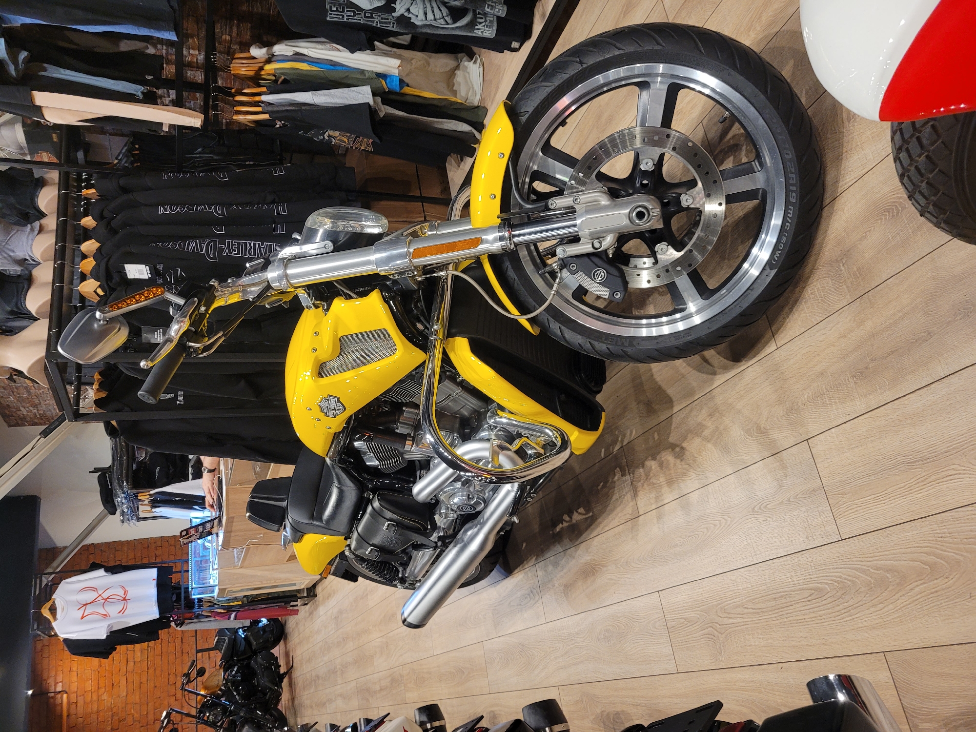 The image depicts a yellow motorcycle, possibly a Harley-Davidson, displayed inside a retail store. Clothing racks with garments are visible in the background, along with other motorcycles. The store appears to have wood-grain flooring. The location is not determinable.