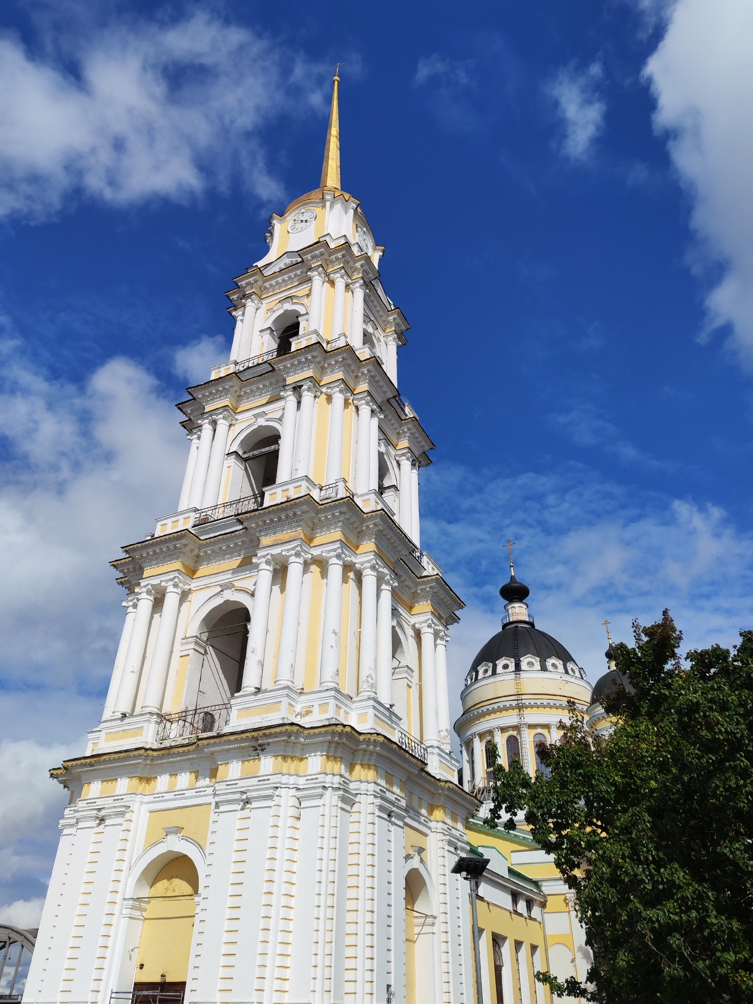 The image shows the bell tower and domes of a church, likely a cathedral, in Bol'shie Mkhi, Russia. The bell tower is tall and slender, painted white with yellow accents. It features multiple levels with arched openings and decorative columns. At the very top, a golden spire rises towards a bright blue sky dotted with white clouds. To the right of the bell tower, two black domes, also characteristic of Russian Orthodox churches, are visible, topped with crosses. The domes are situated on a building with yellow and white architectural elements. A lush green tree partially obscures the right side of the scene. The perspective is low-angle, looking up at the structures against the sky. There are no people or animals visible in the image.