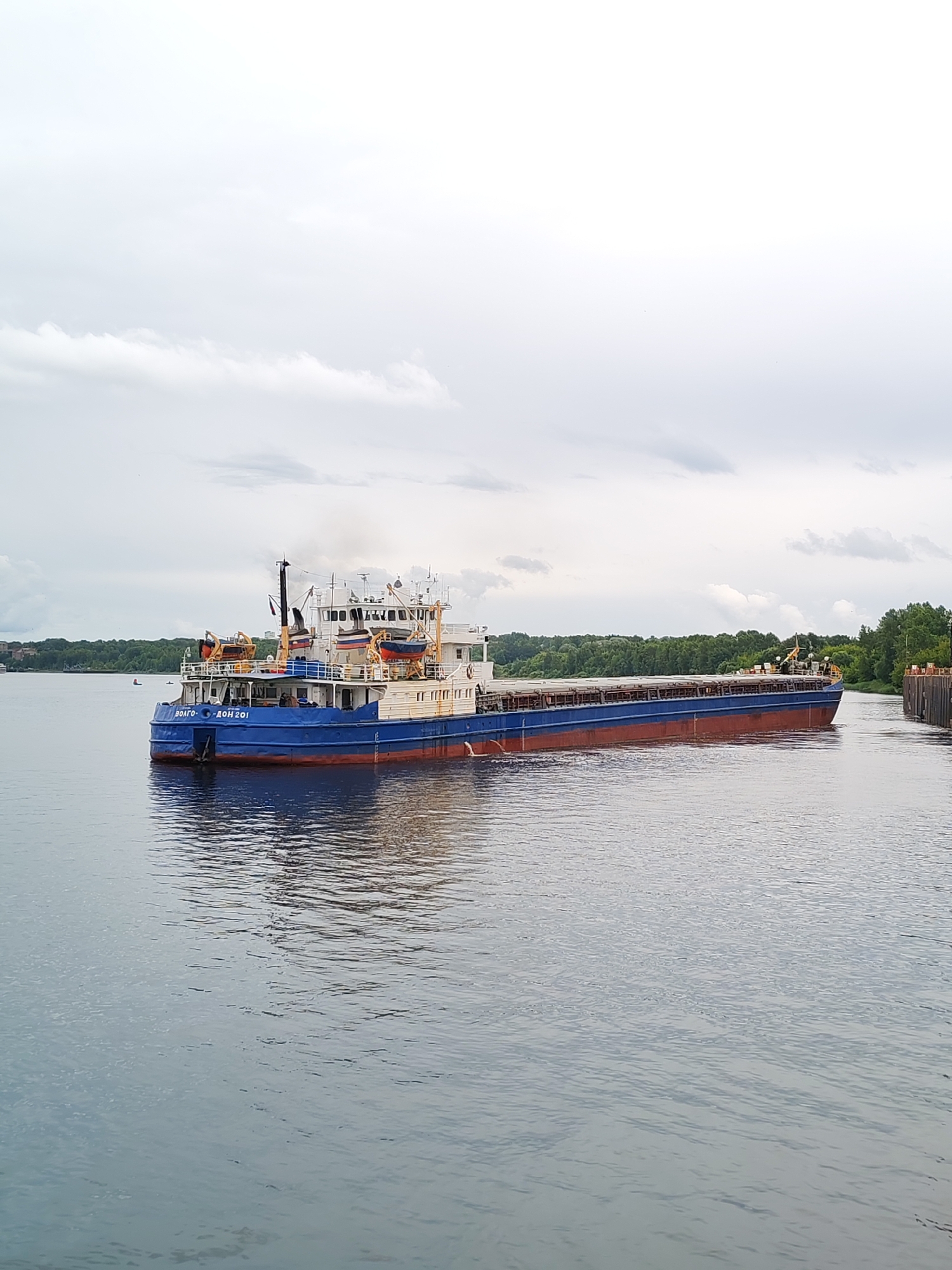 A barge labeled "ВОЛГО-ДОН 201" is traveling on a body of water, likely a river, in Bol'shie Mkhi, Russia. The sky is overcast with gray clouds. The barge is blue and red with white superstructures. It appears to be carrying cargo under a tarp. There are trees and structures visible on the distant shore.