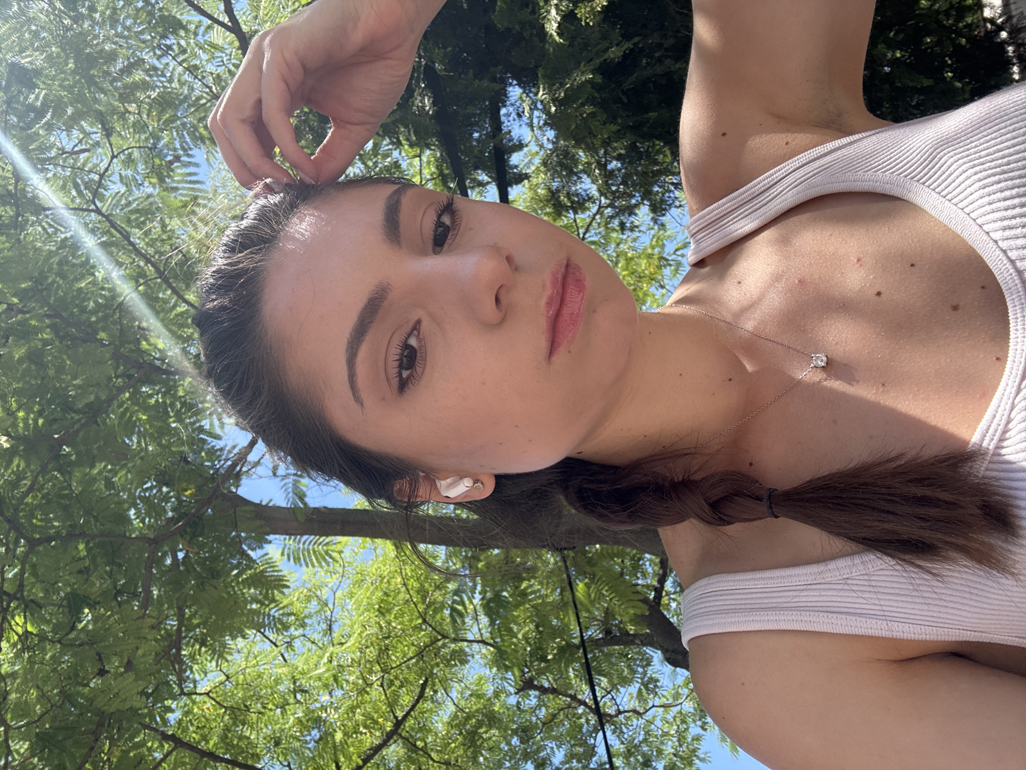 A young woman with dark brown hair in a braid, wearing a light pink ribbed tank top and a silver necklace with a diamond pendant, is pictured outdoors. She has earbuds in her ears and is touching her hair with her right hand. The background consists of green trees and a blue sky, suggesting a sunny day. The image appears to be a selfie taken from a low angle. There is no identifiable information or context provided that would allow for the determination of a specific location in Varna, Bulgaria.
