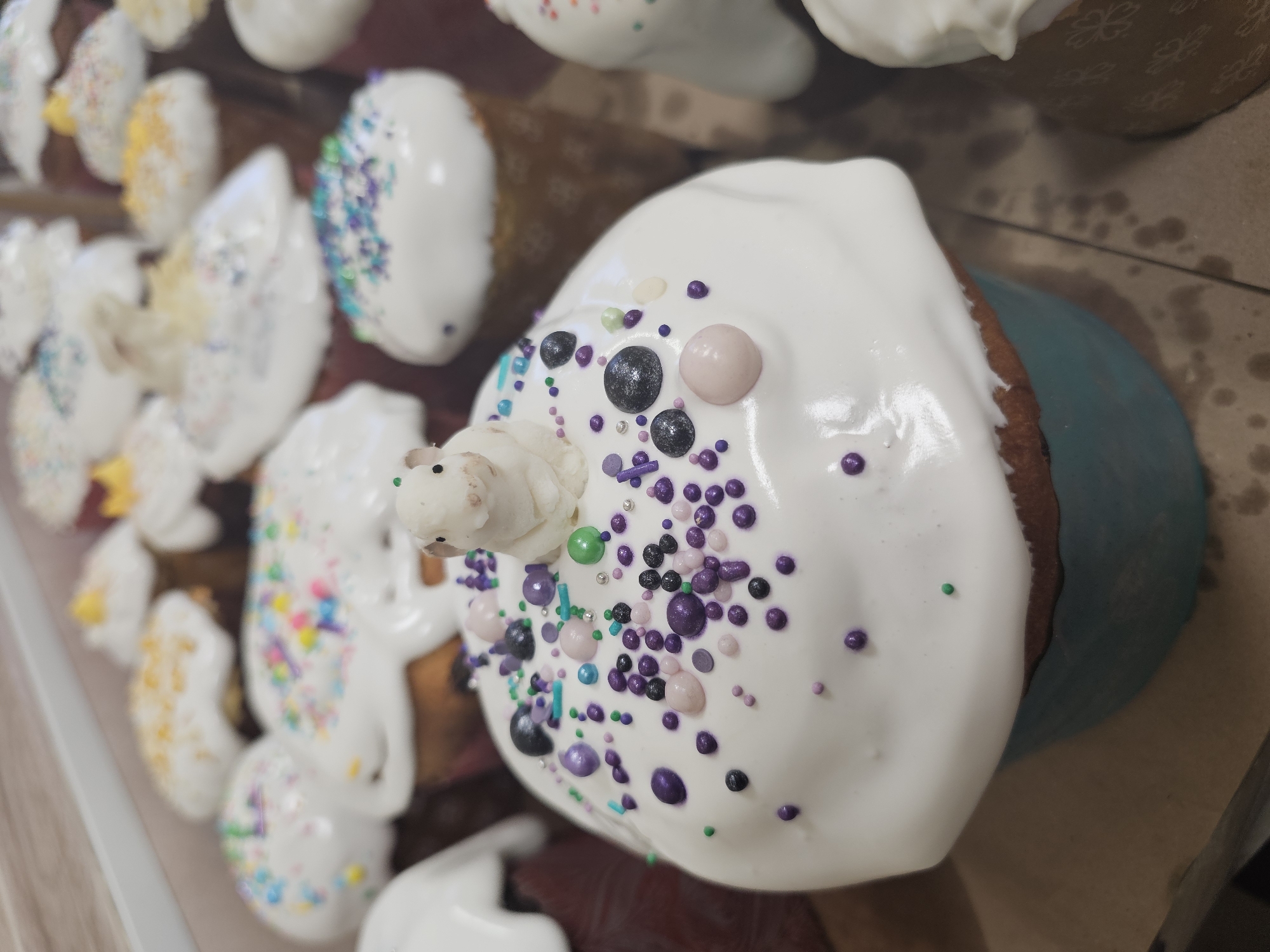 The image depicts a close-up of several decorated Easter cakes (kulich) likely displayed for sale in a shop or bakery. The cakes are topped with white icing and various colorful sprinkles and edible decorations. The main cake in the foreground features a small, white sugar figurine of a lamb or other animal. The scene is probably in Moscow, Russia due to the popularity of decorated kulich cakes for Easter celebrations in the Russian Orthodox tradition.
