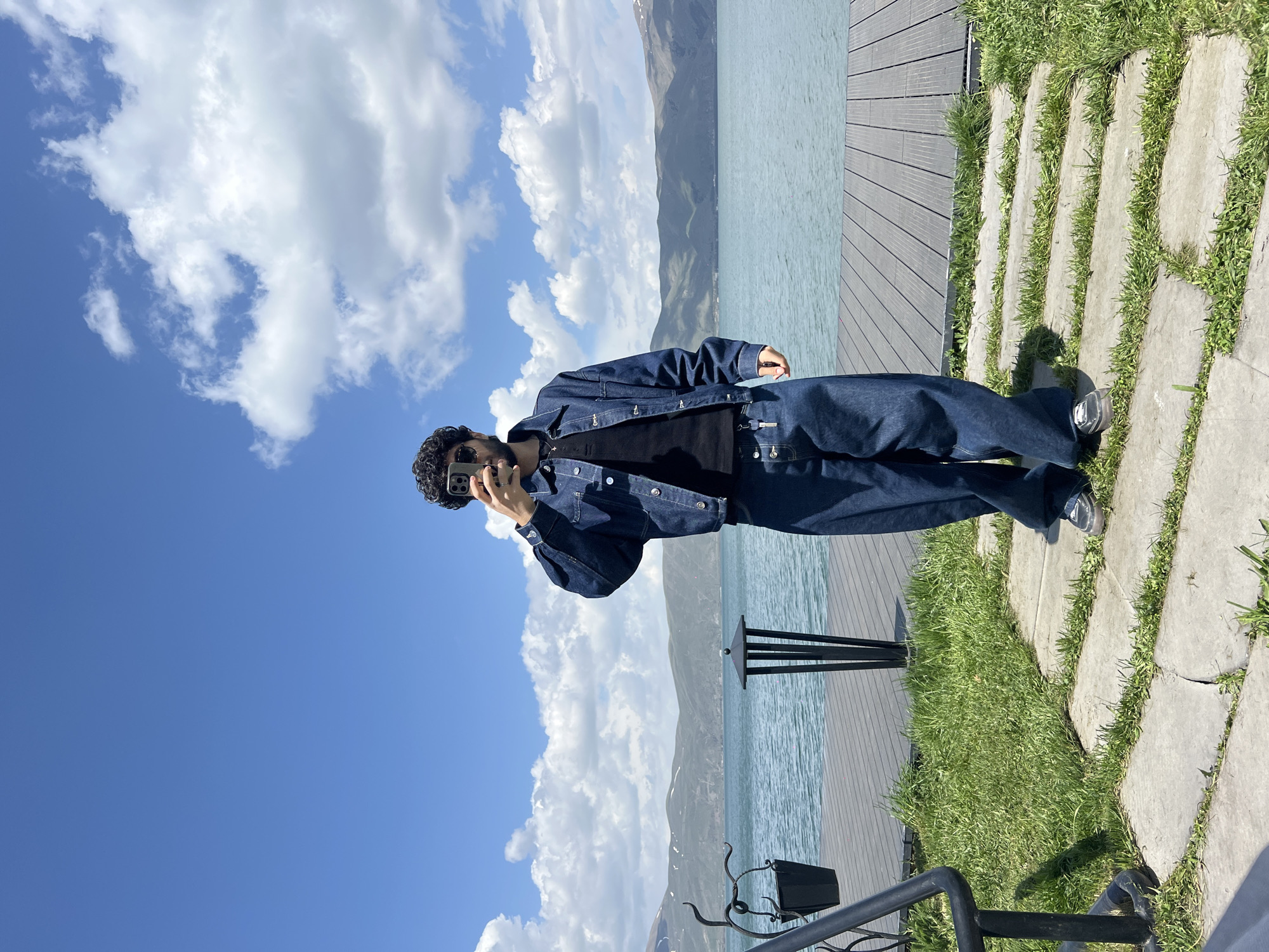 A person dressed in a dark denim jacket and pants stands on a wooden platform next to a body of water, possibly a lake, with mountains in the background, under a sky with scattered clouds. The person is holding a phone to their face, possibly taking a picture. There's grass and stone steps along the edge of the platform, and metal railings are visible in the foreground. The city location is unknown.