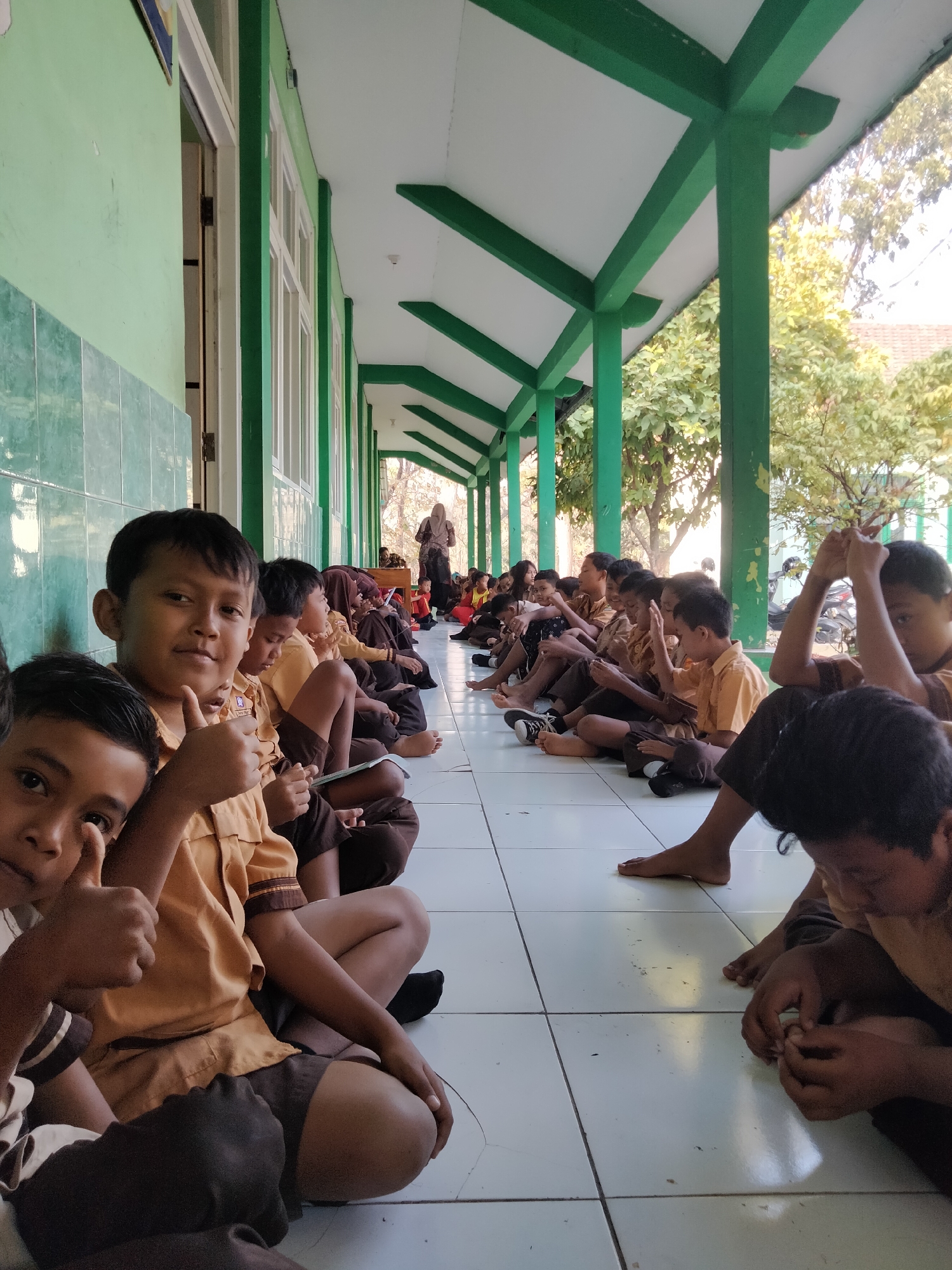 In Pucakwangi, Indonesia, a group of elementary school children, predominantly boys, are seated on the tiled floor of an outdoor school hallway. They are dressed in light brown uniforms with darker brown shorts and socks. The hallway has a green painted wall on the left and a green-painted covered walkway with white ceiling beams extending to the right. Many of the children are looking down, some are holding books or notebooks, and a few are interacting with each other. The child in the foreground on the left is giving a thumbs-up gesture, and the child in the foreground on the right is examining their fingernails. Trees with yellowing leaves are visible beyond the covered walkway, suggesting it might be late in the dry season or a time of year when these trees shed leaves. Sunlight is casting shadows, indicating it is daytime. A woman in a headscarf and long dress stands further down the hallway, appearing to address the children.