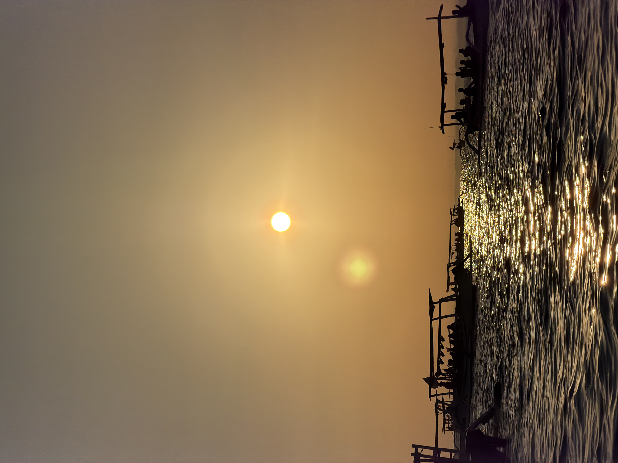 The image depicts a serene sunset over the waters of Buleleng, Indonesia. Several traditional Balinese boats, known as jukung, are silhouetted against the warm, golden sky. These boats are filled with passengers, their figures indistinct due to the backlight. The sun, a bright orb, is low on the horizon, casting a shimmering path of light across the gentle waves. The overall atmosphere is tranquil and picturesque, characteristic of a sunset in this coastal region.