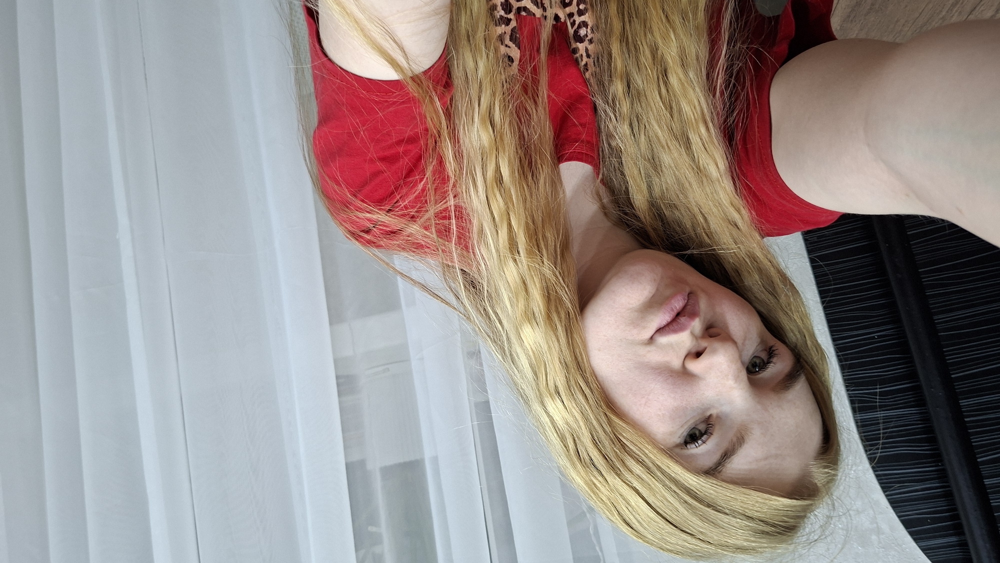 An image, captured from a slightly elevated and tilted perspective, features a young woman with long, wavy blonde hair cascading around her shoulders. She wears a red short-sleeved top, with a glimpse of a leopard print garment visible underneath near her left shoulder. Her fair skin shows a subtle flush on her cheeks. She has light-colored eyes and looks directly at the viewer with a neutral, slightly pouting expression; her lips are a natural pink.
The background primarily consists of sheer, white or light grey curtains, suggesting an indoor setting, likely near a window. To the right of the woman, a dark, textured surface, possibly a piece of furniture, and an adjacent lighter surface are visible. The scene is illuminated by bright, natural light, indicating it is daytime. No text is visible in the image. The general location context is Chaikovsky, Russia.