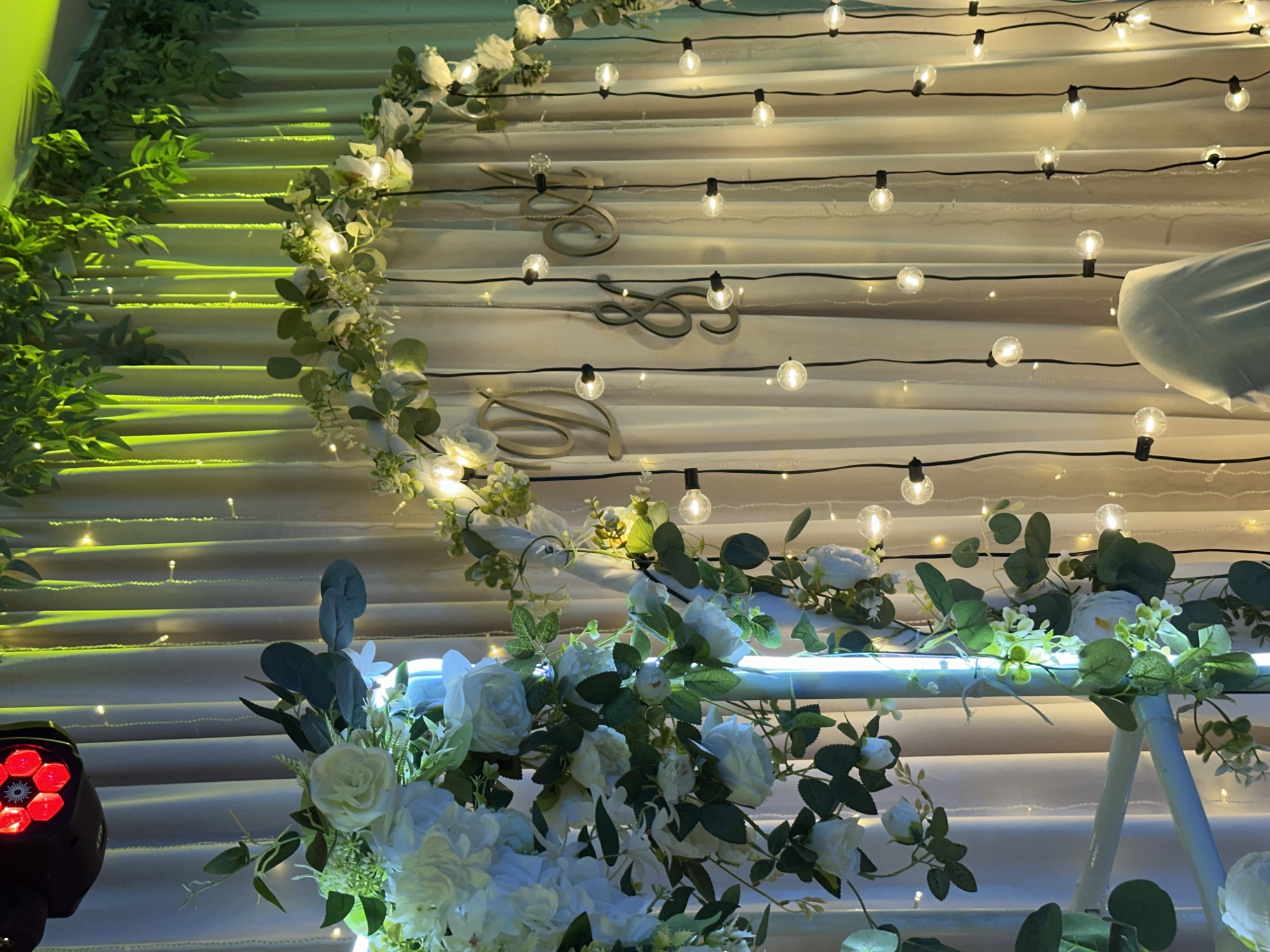 The image depicts a decorated event backdrop, likely for a wedding or special occasion. A beige curtain serves as the primary background, illuminated by strings of warm fairy lights and overhead lighting that casts a green hue. On the left, a prominent floral arrangement features white roses and greenery cascading down. A vertical, illuminated blue light fixture is partially visible. In the center, a large, ornate ampersand symbol is affixed to the curtain, flanked by wooden letters that appear to spell "D & M". Several strings of Edison-style bulbs hang vertically in front of the curtain, some with visible black cords and holders. A partially covered white chair is visible in the lower right corner. The overall ambiance is festive and romantic, suggesting an indoor event space. The image does not provide specific details or landmarks that would allow for definitive placement within Chaikovsky, Russia.